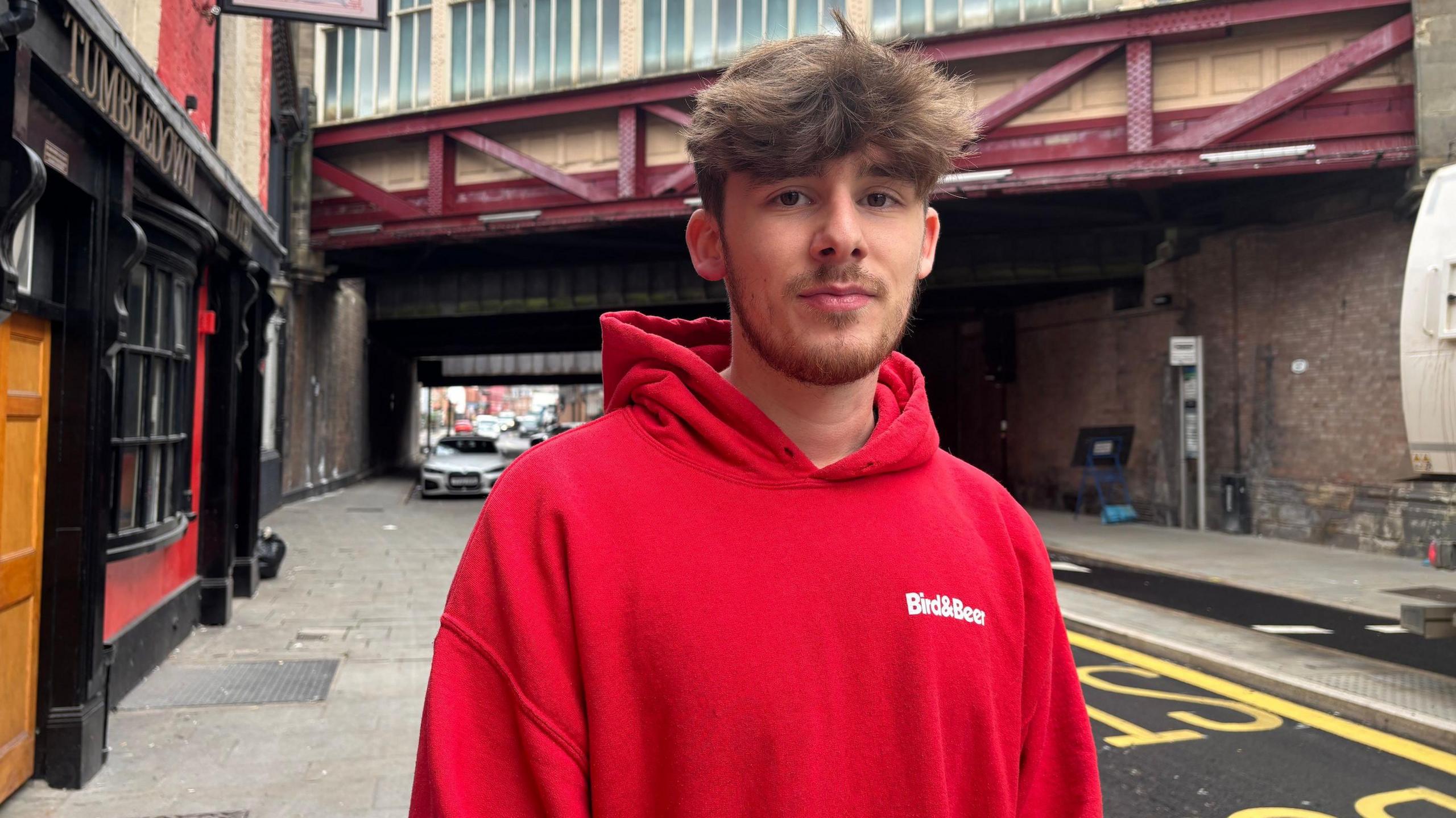 A man with brown hair, wearing a red hoodie with a logo that says Bird & Beer, is standing on a narrow road in front of a bridge.