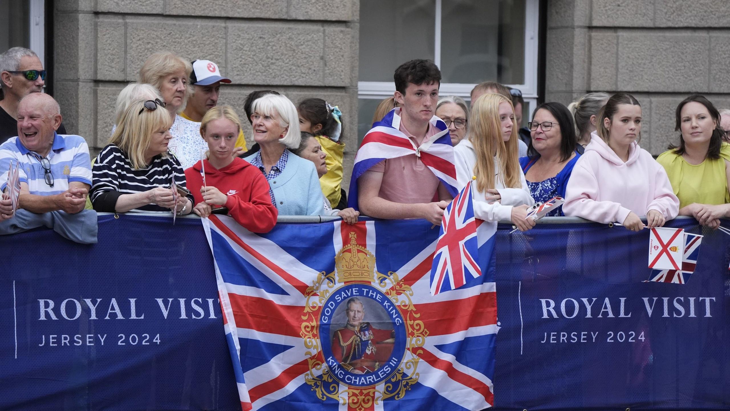 A crowd of people, many holding Union Flags and Jersey flags, wait behind barriers for the King and Queen.