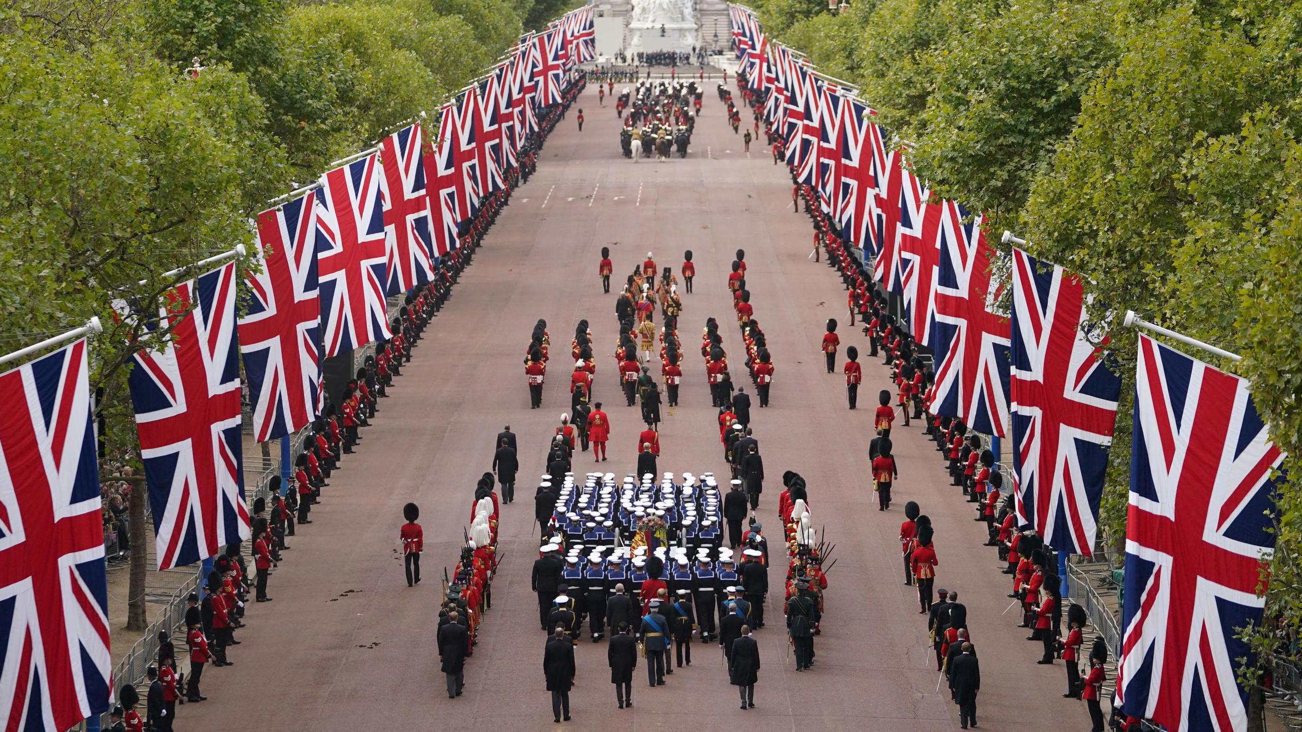 A long street is lined with flags. Ceremonial soldiers march up the street around a coffin. 