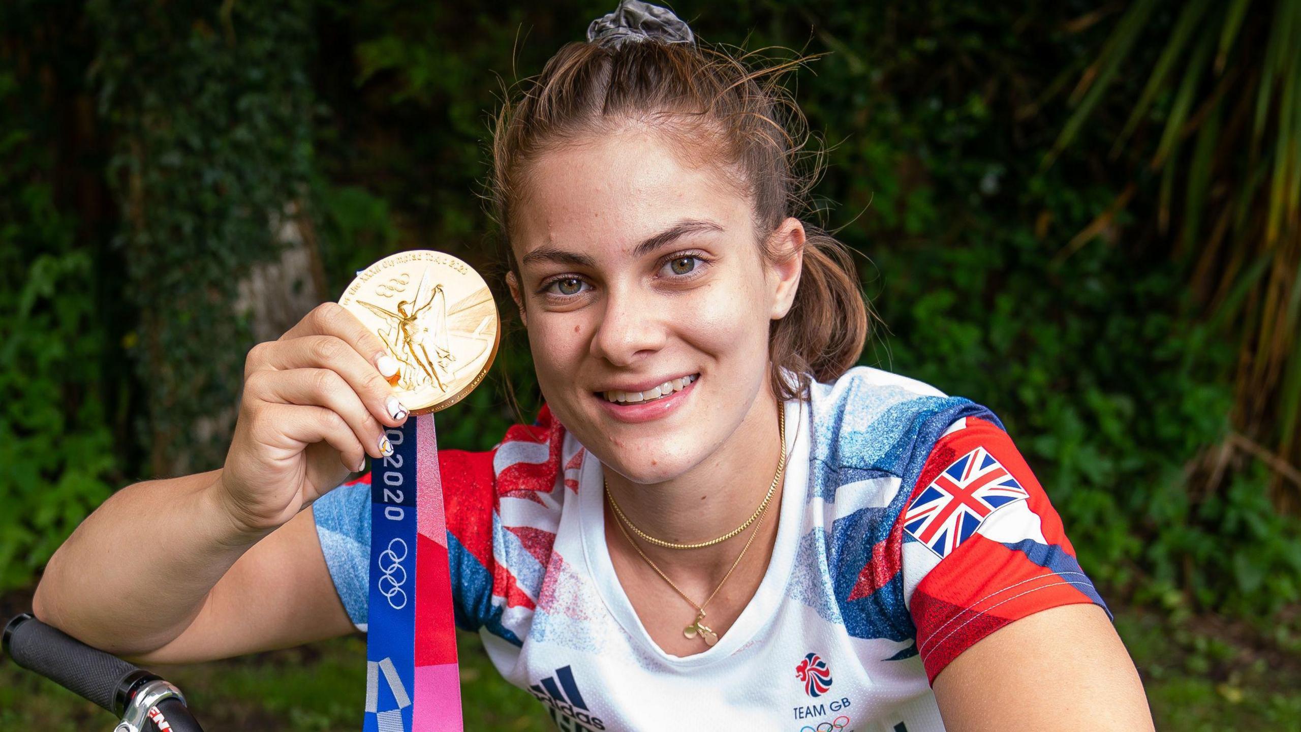 A woman holding a medal while resting her elbow on her bike. She has her hair in a ponytail and is smiling at the camera 