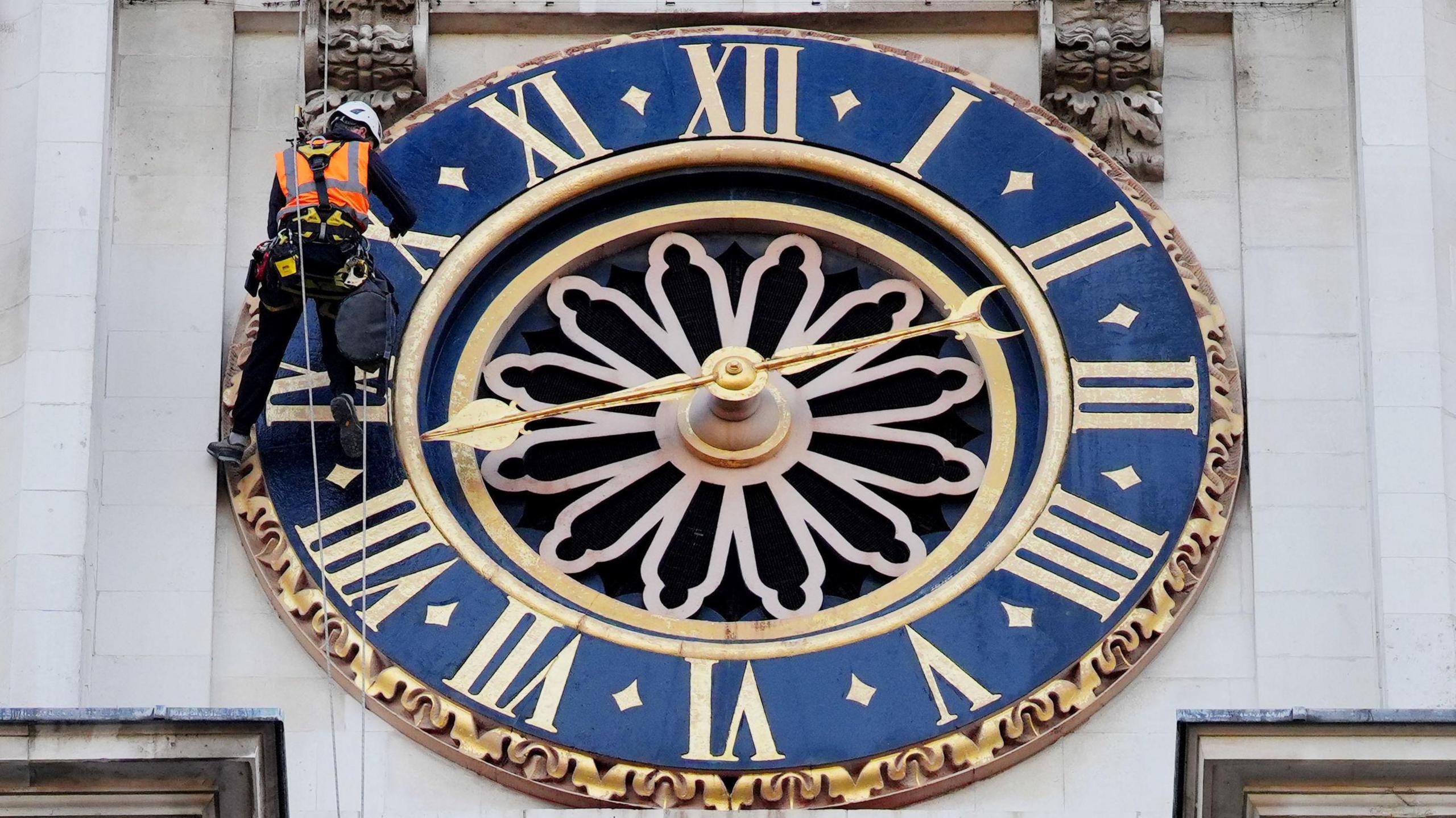 A man in high-visibility wear surveys a blue and gold clock face on the Hawksmoor clock tower at Westminster Abbey.