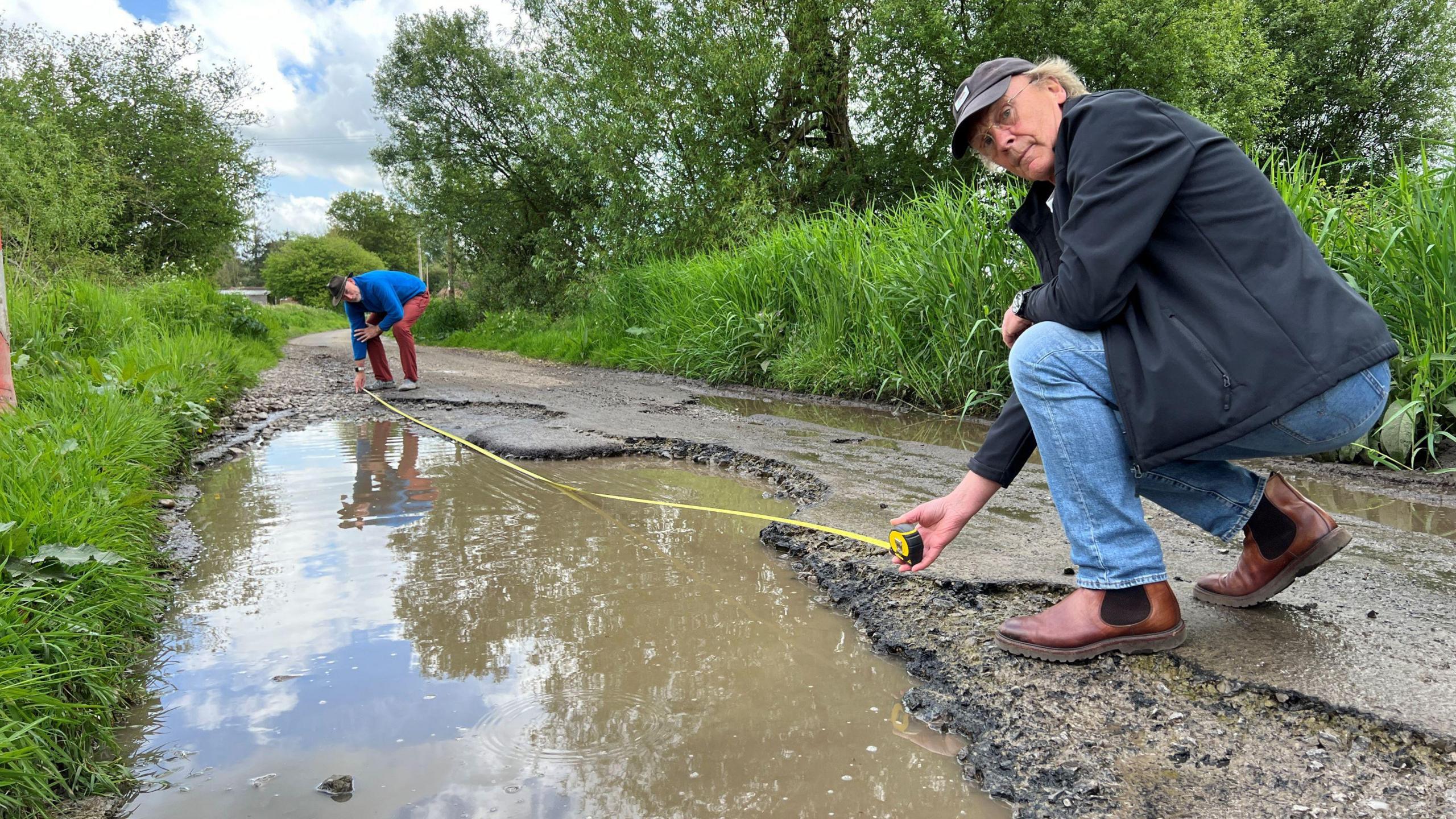 Two men holding a tape measure of a large pothole which is flooded