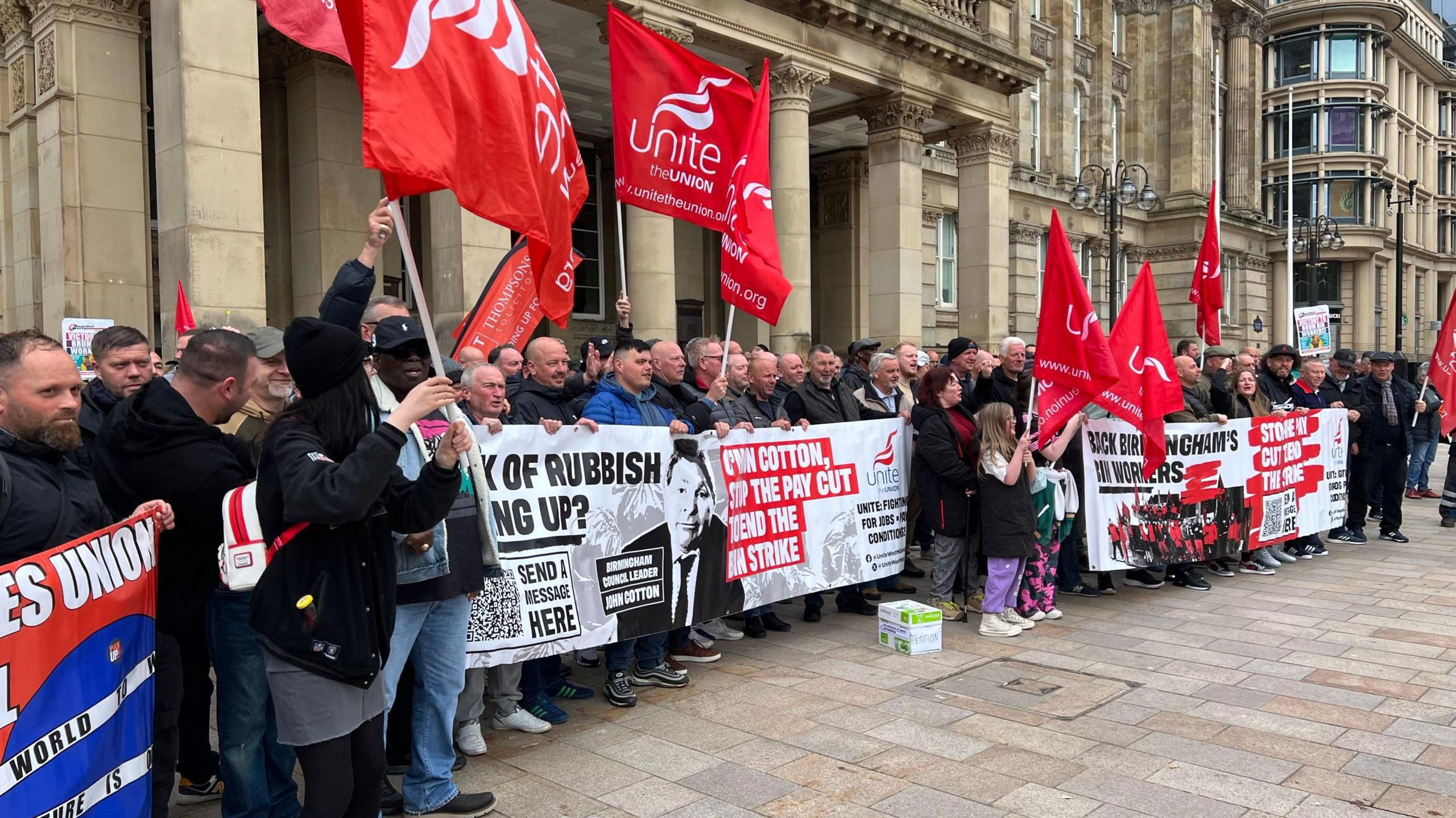 A group of people stand in a row and hold red flags and banners in different colours. The red flags say "Unite the union" on them in white writing. The crowd mostly contains men but there are a few women also.