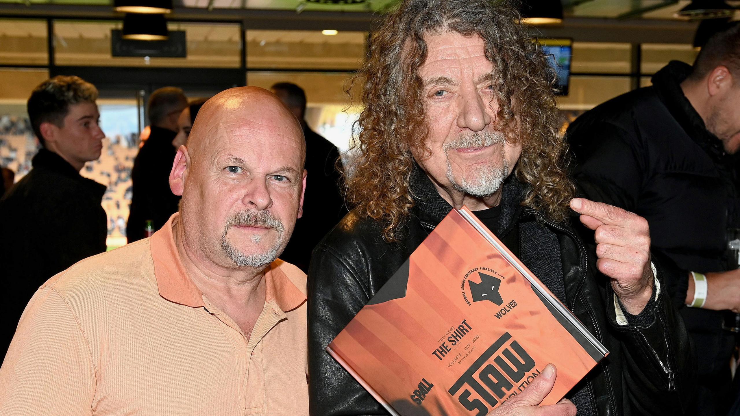 Two men, one holding a coffee table book about Wolves football shirts. A bald man is wearing an orange polo shirt. The other man has long curly hair, a goatee beard and is wearing a black leather jacket