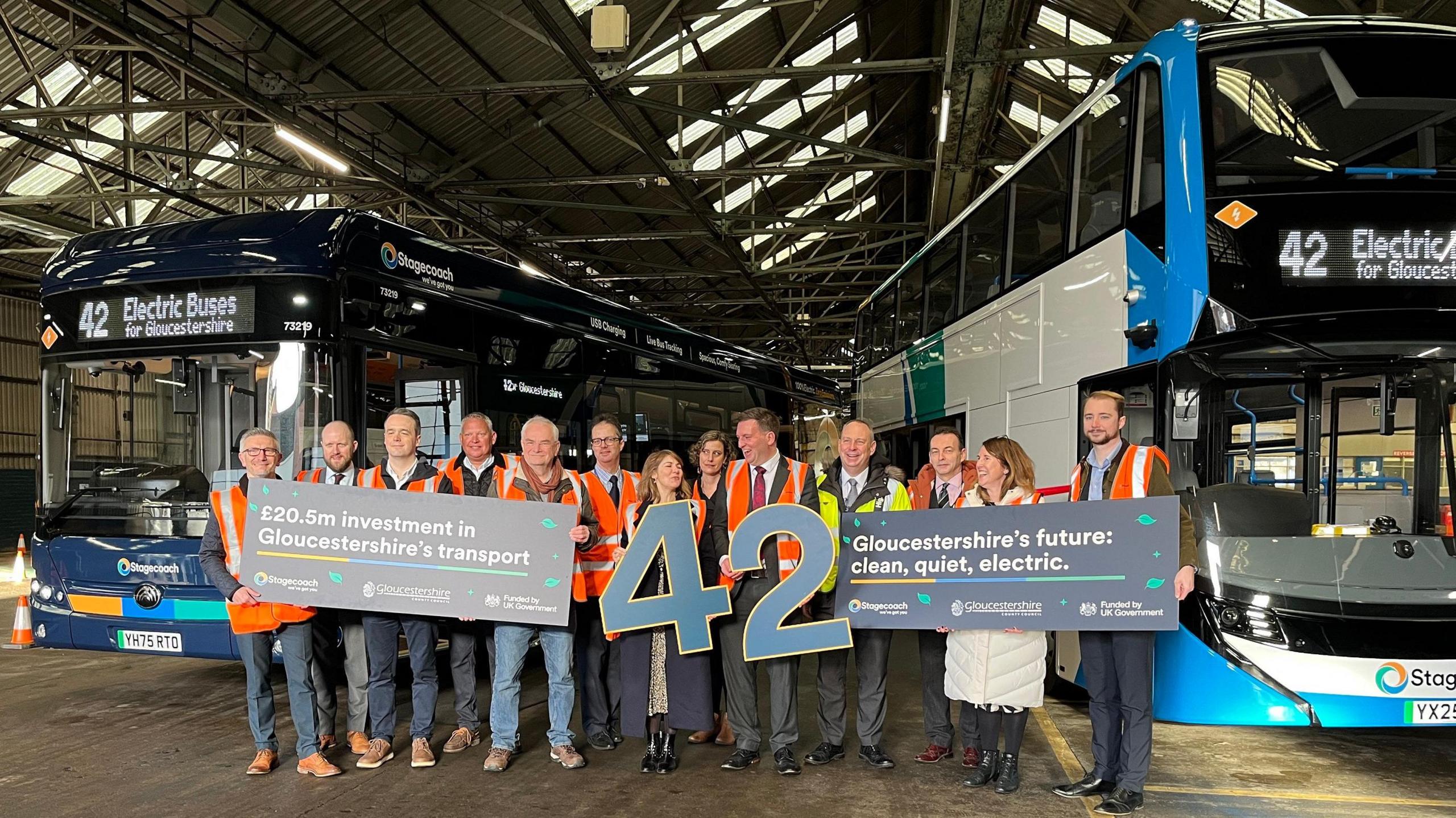 One single deck and one double decker bus inside a large open building (the depot). Several Stagecoach West workers stand before the buses, holding two large blue numbers that read 42. They are also holding signs with one saying "£20.5m investment in Gloucestershire's transport" and the other saying "Gloucestershire's future: clean, quiet, electric".