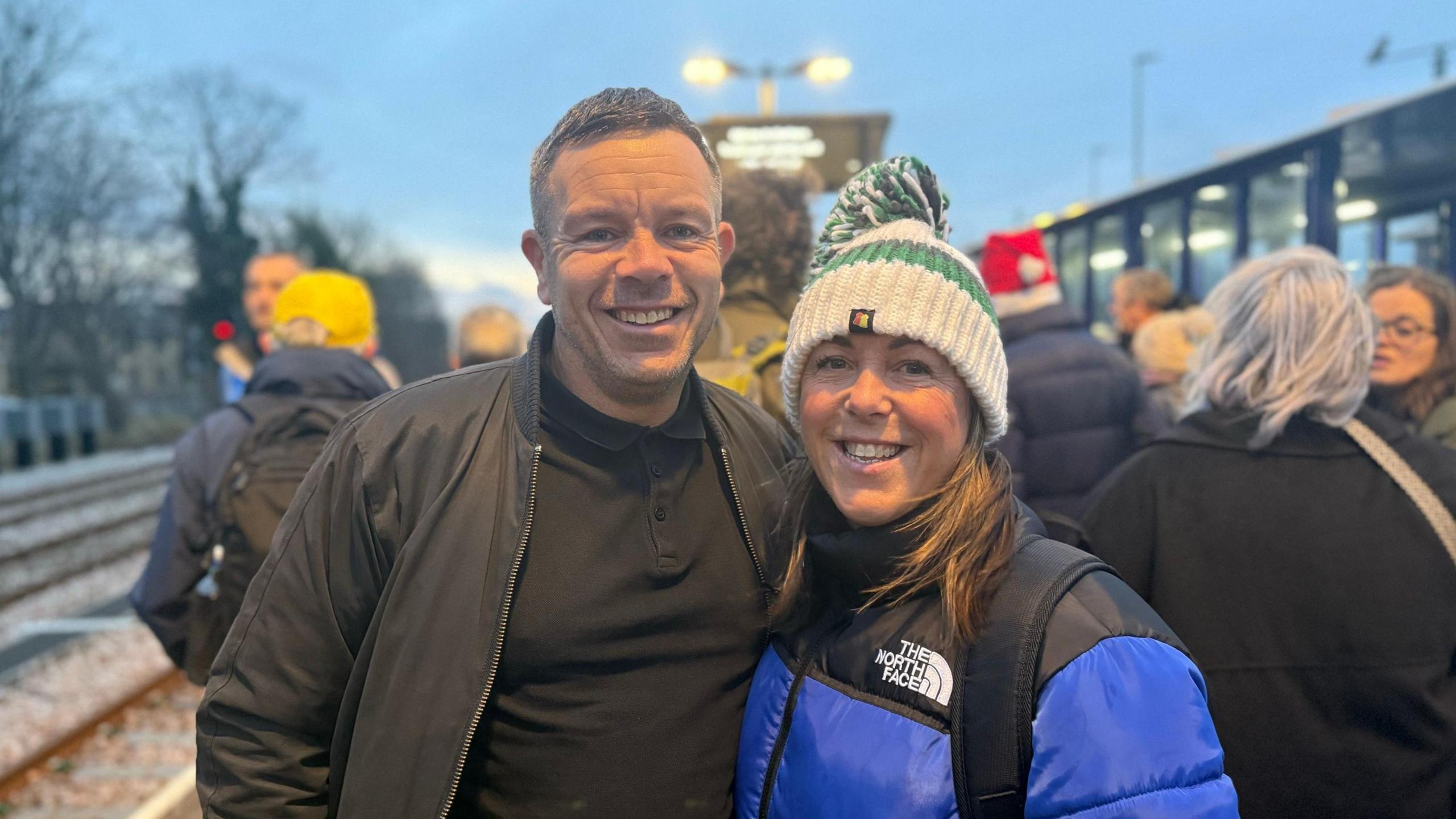 Kelly Kay has a green and white bobble hat and a blue jacket. She is standing next to a man wearing a black top and a black jacket. 