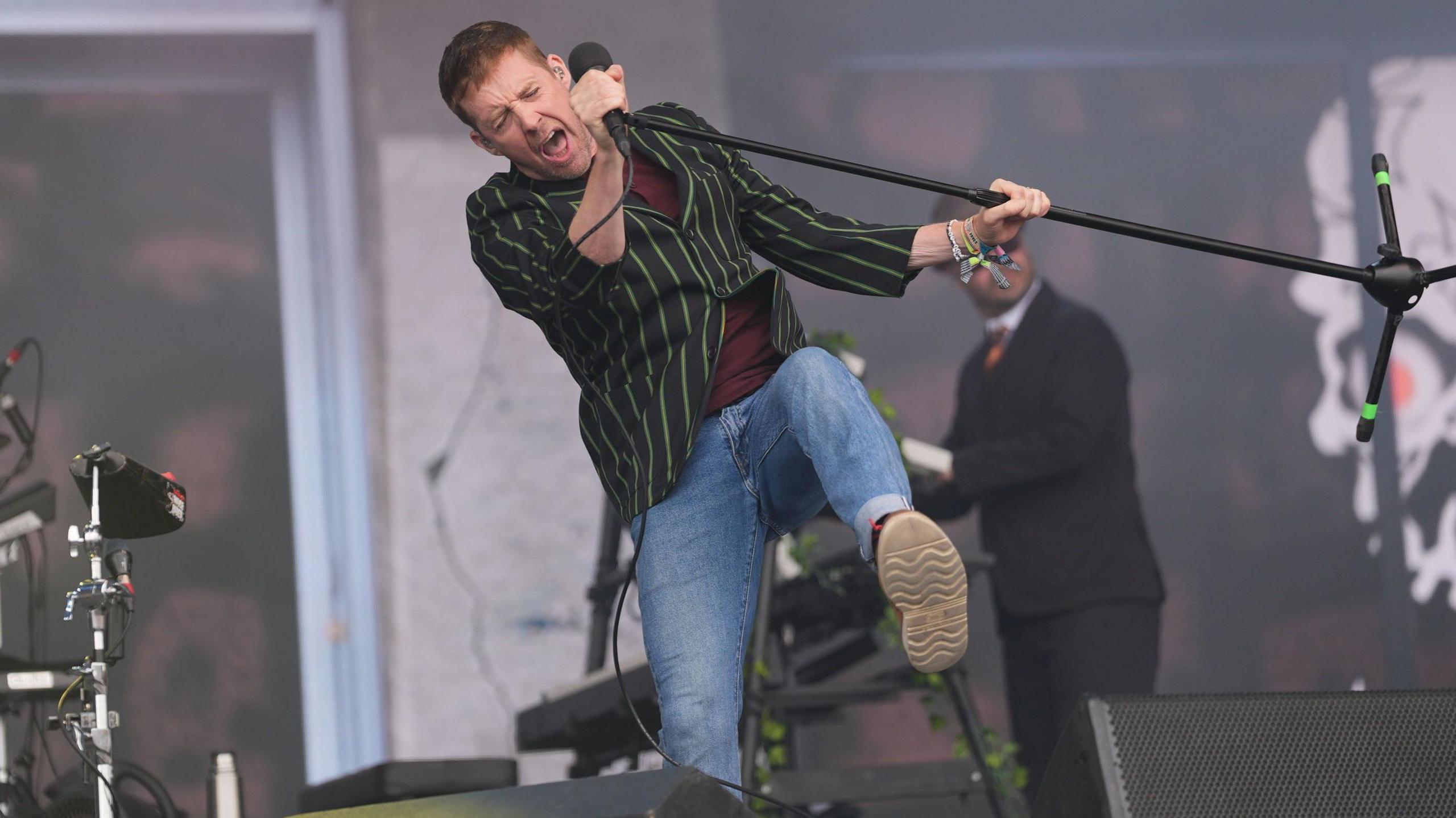 Ricky Wilson sings into a microphone as he kicks his foot up in the air on a stage.