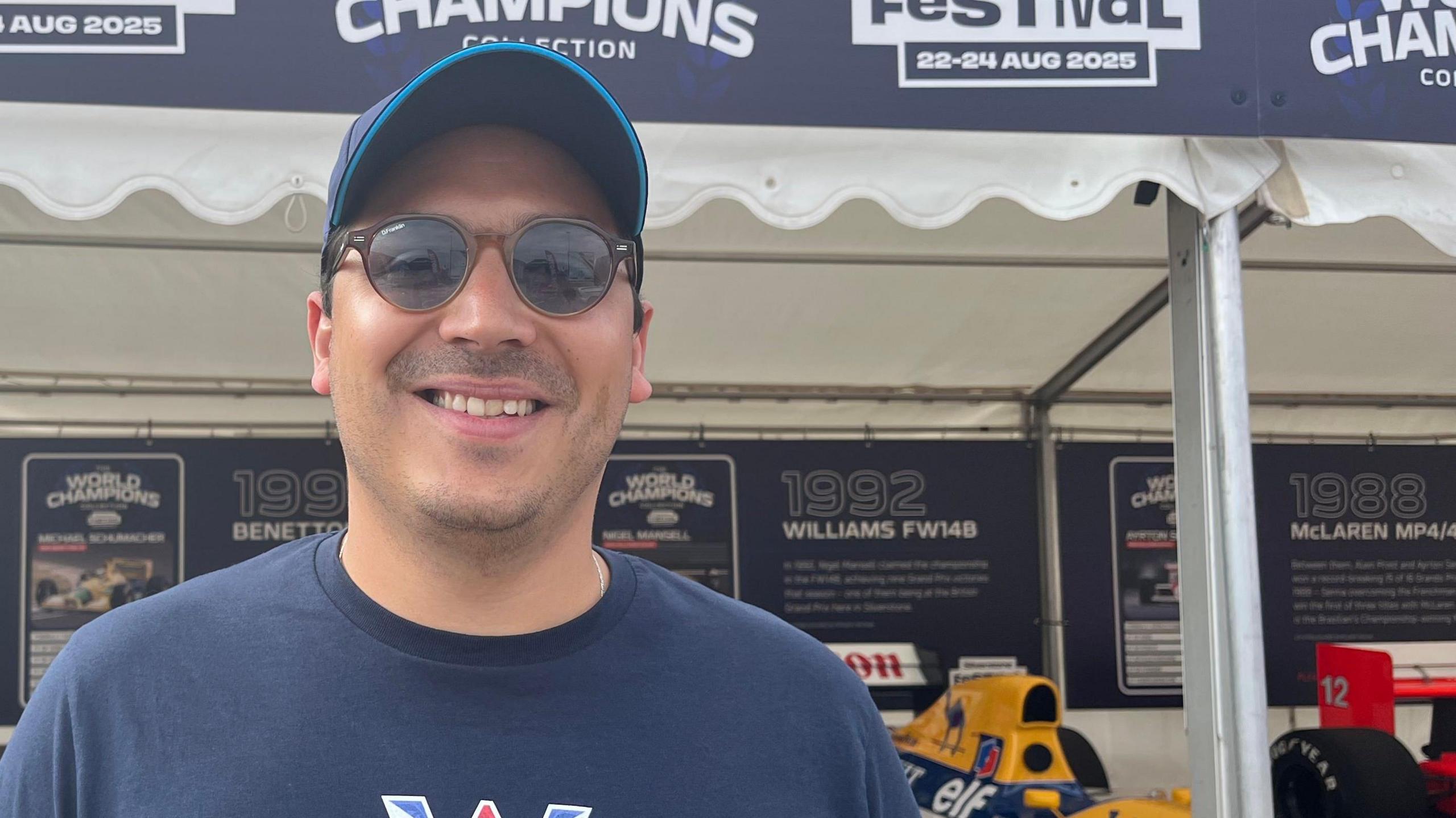 A man wearing a navy blue Williams race team T-shirt and cap with sunglasses on is smiling at the camera. Behind him is a collection of formula one cars, with a yellow Williams FW148 car from 1992 slightly visible.