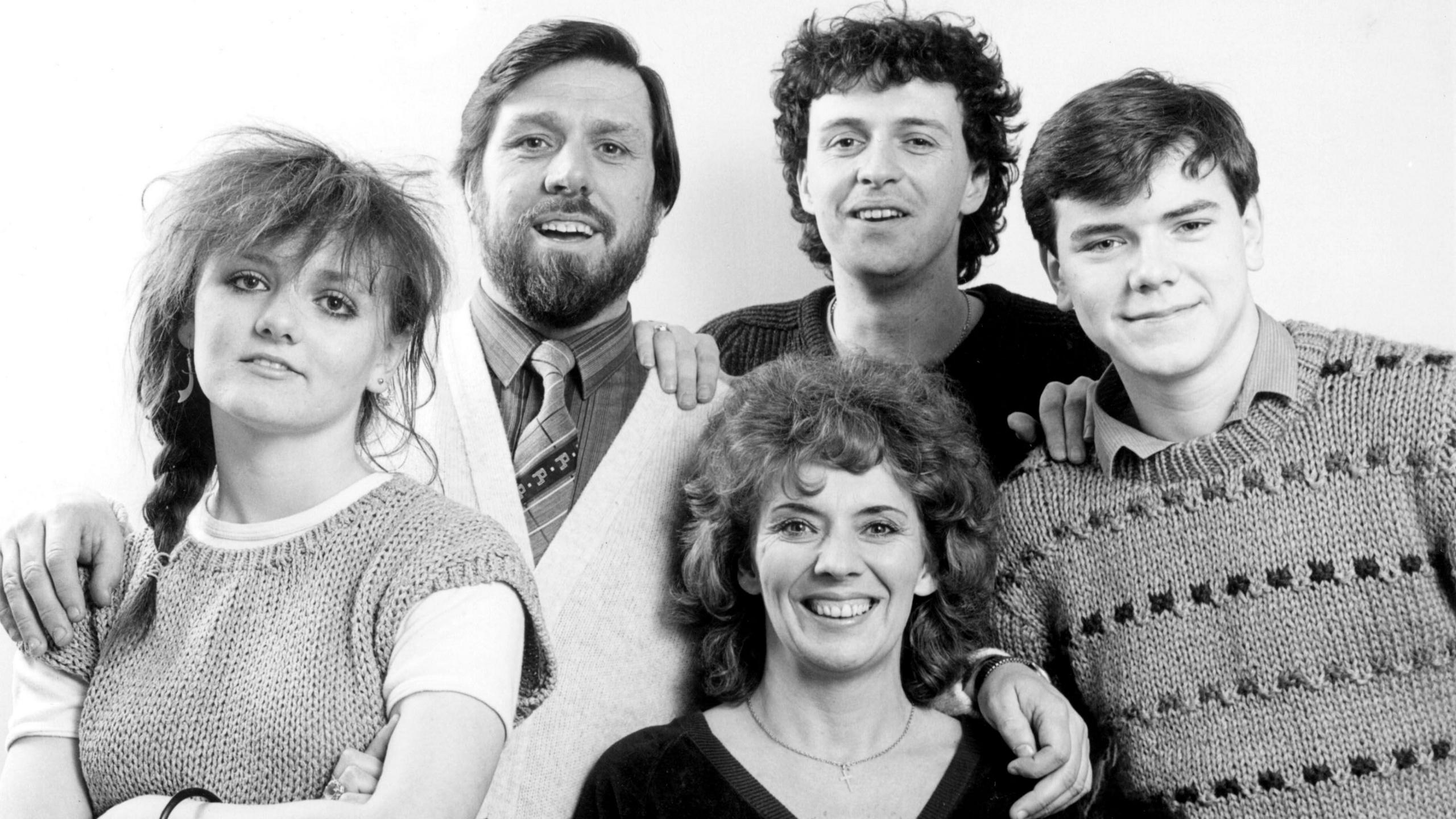 A black and white photograph of the Grant family, featuring parents Bobby, portrayed by Ricky Tomlinson, and Sheila Grant, played by Sue Johnston, along with their children Barry, Karen and Damon. They are all smiling at the camera and have their arms around each other.