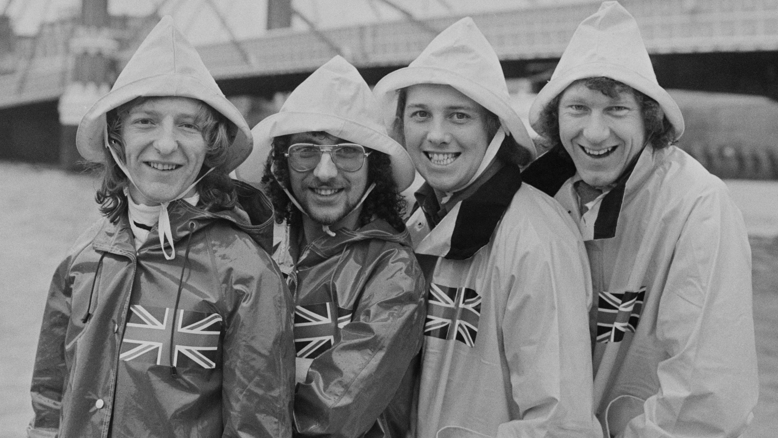 British pop band Black Lace posing wearing raincoats with union jack flag on front and rain hats on the River Thames bank near Albert Bridge, London, UK, 9th March 1979