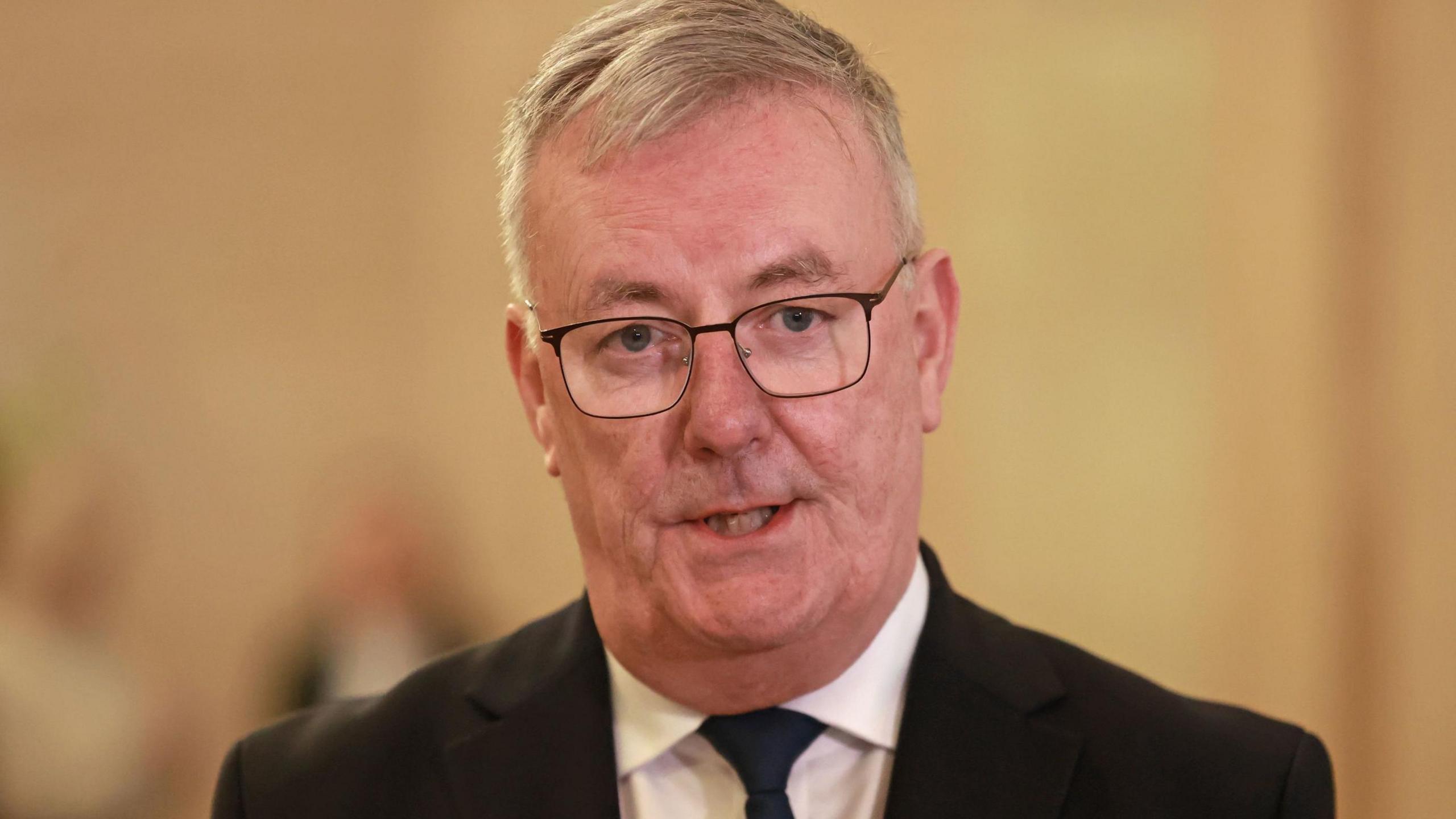 Mike Nesbitt - He has grey hair and is wearing a black suit, black tie and white shirt and black-rimmed glasses which are rectangular in shape.