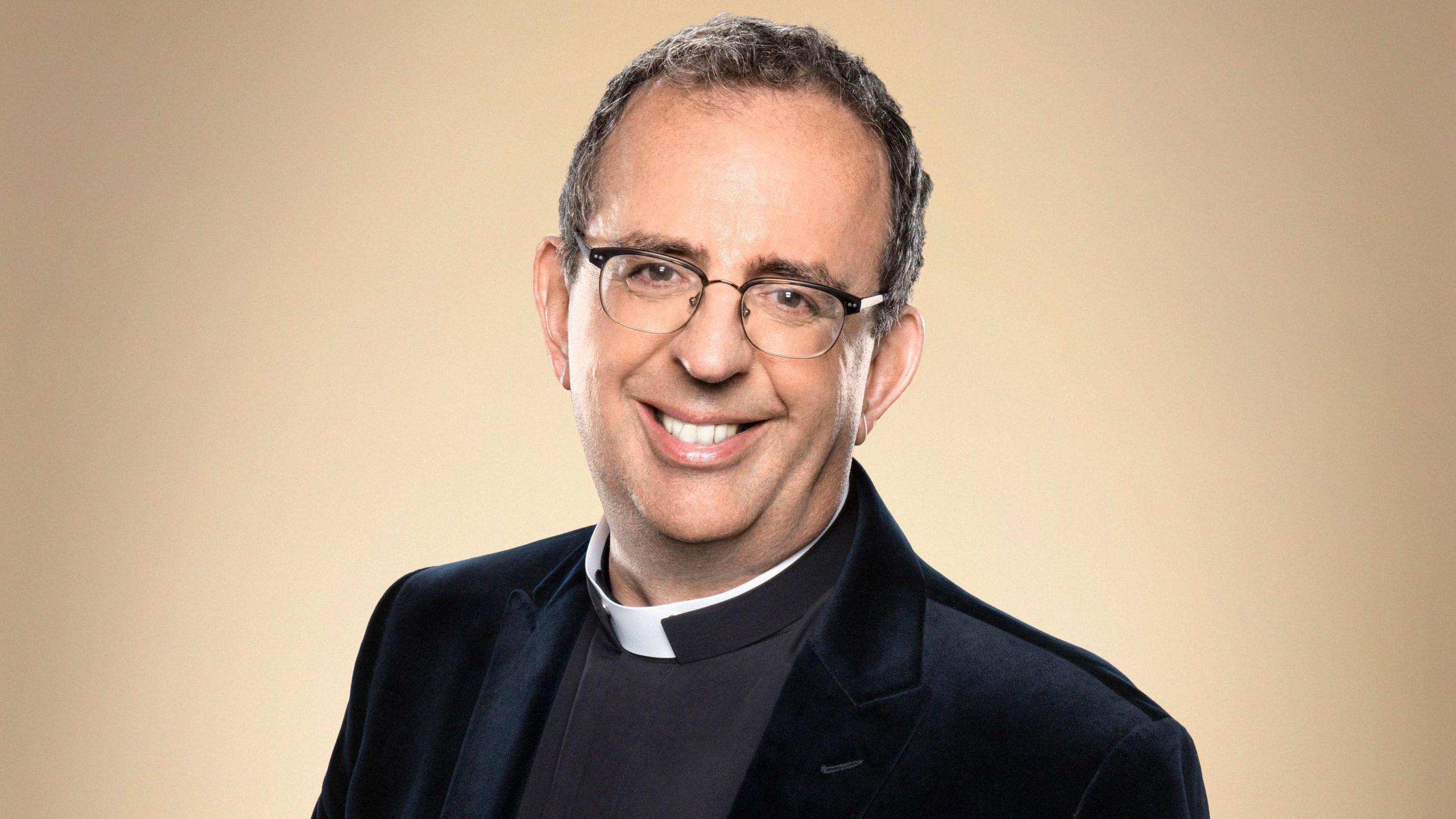 Rev Richard Coles wears a clerical collar and black blazer while smiling directly into the camera.