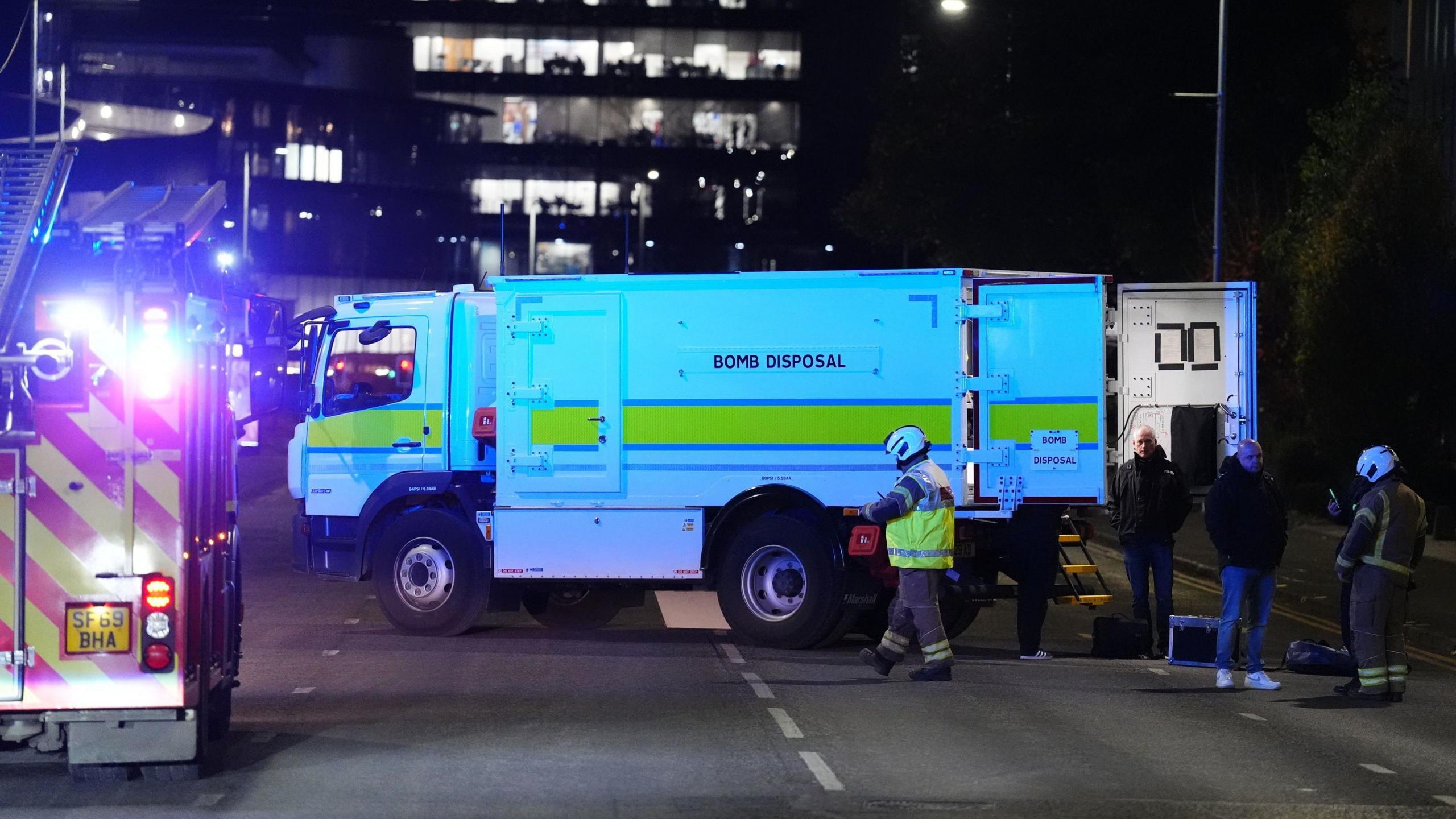 A bomb squad van and fire engine blocks the road close to the bus station.   It is night and the vans have their lights on. Fire figheters walk arounf the vehicles.