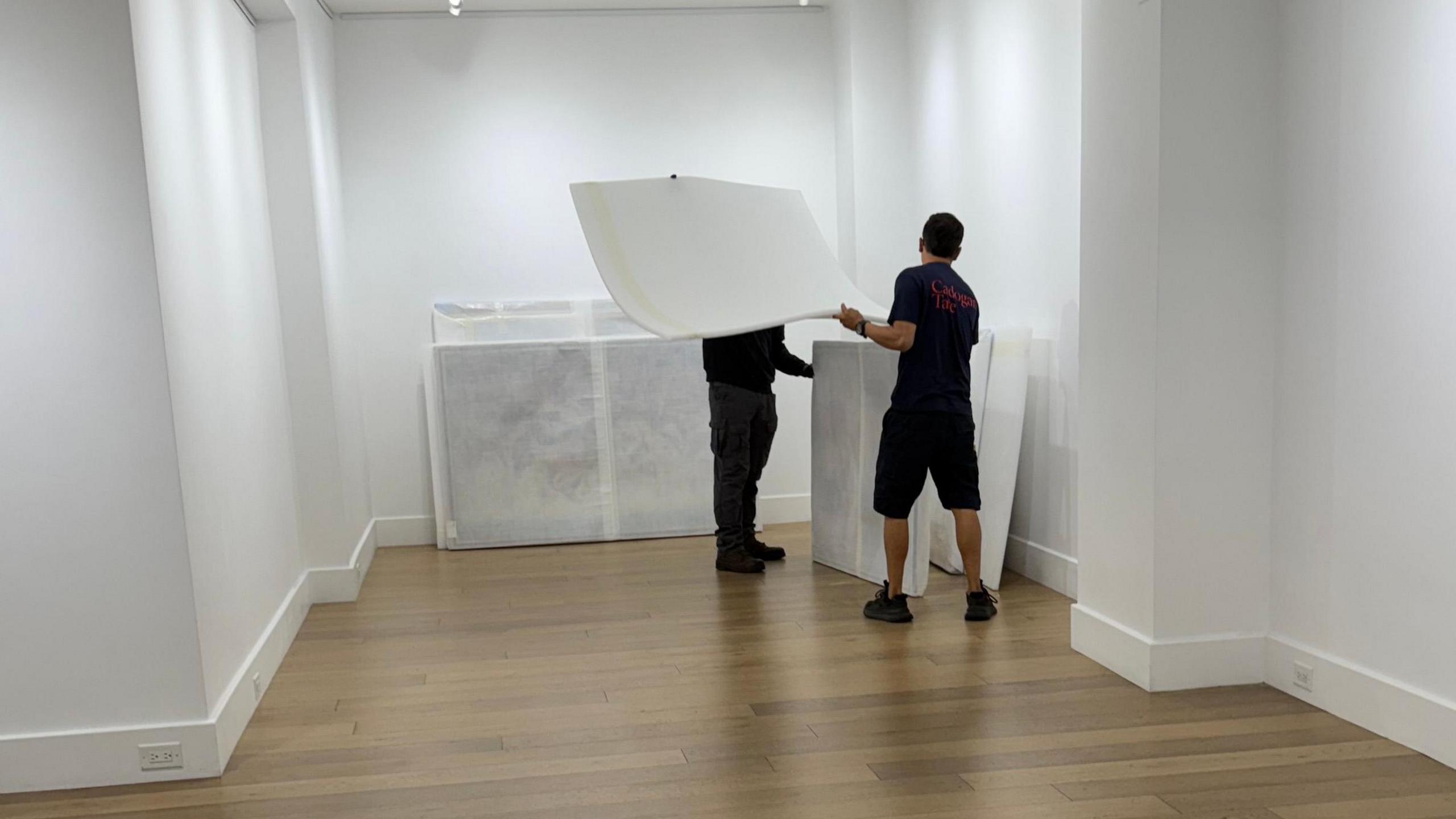 Two people beside wrapped up canvases leaning against white walls. They are removing foam
