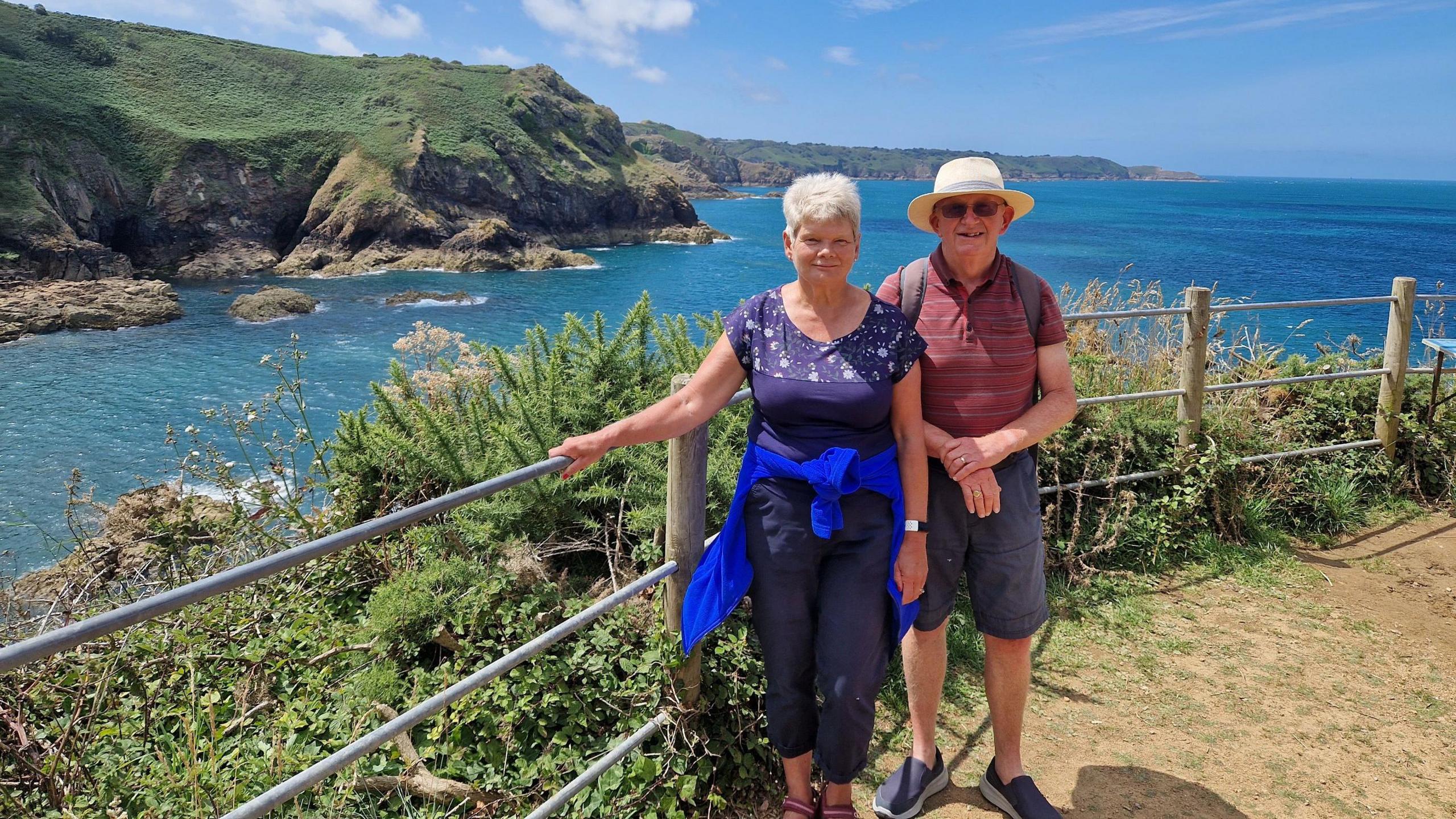 Steve and Vanessa Butcher smile at the camera wearing summer clothes and holding onto a fence overlooking the sea and cliffs on a sunny day