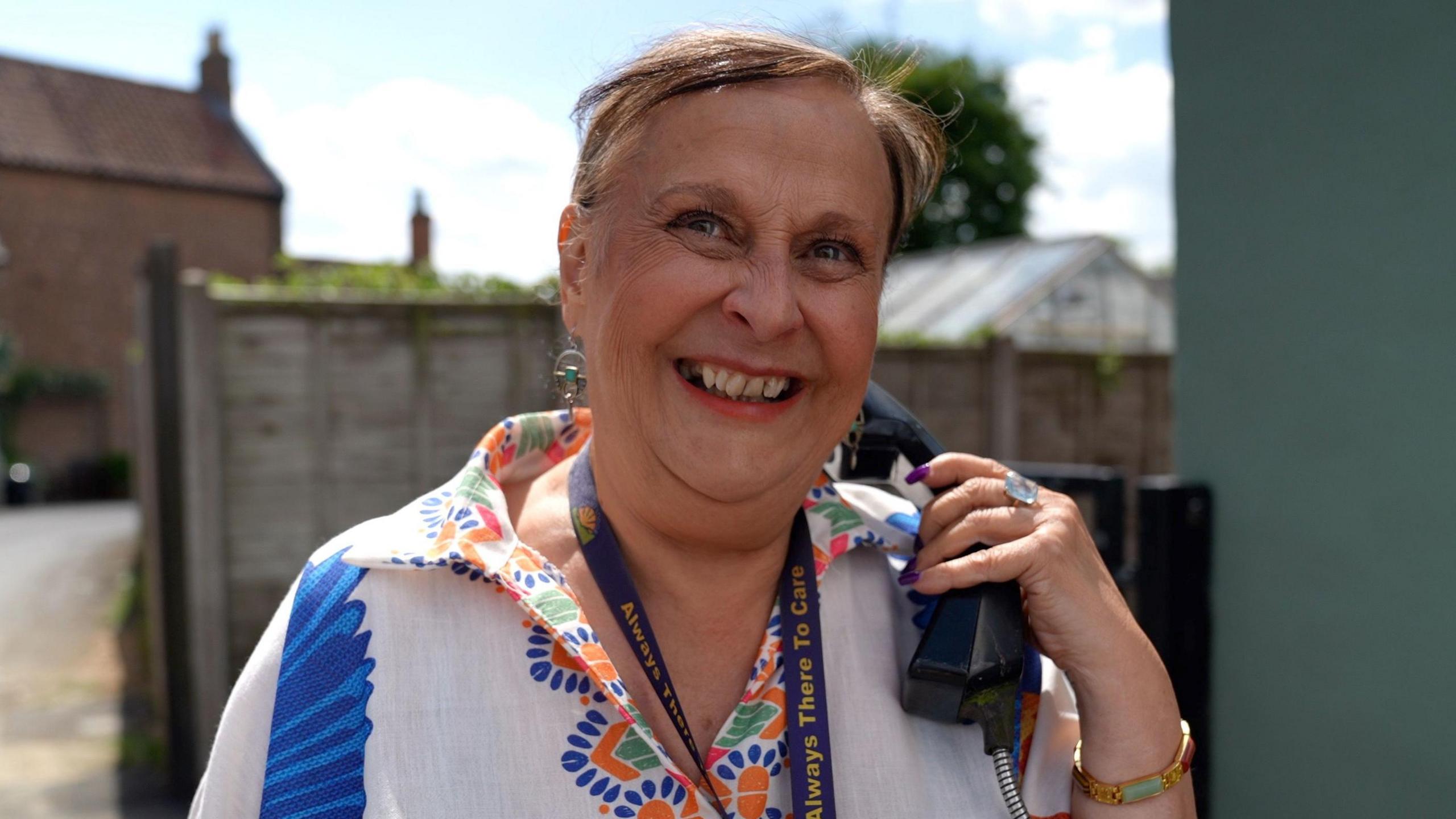 Caroline Ali, a middle-aged woman, is smiling at the camera while holding a black payphone in her left hand up to her ear. She has long purple nails, red lipstick on and is wearing a white blouse with a colourful pattern on the collar. She has brown hair tied back and is wearing a gold bracelet and a large sparkly ring on her left hand.