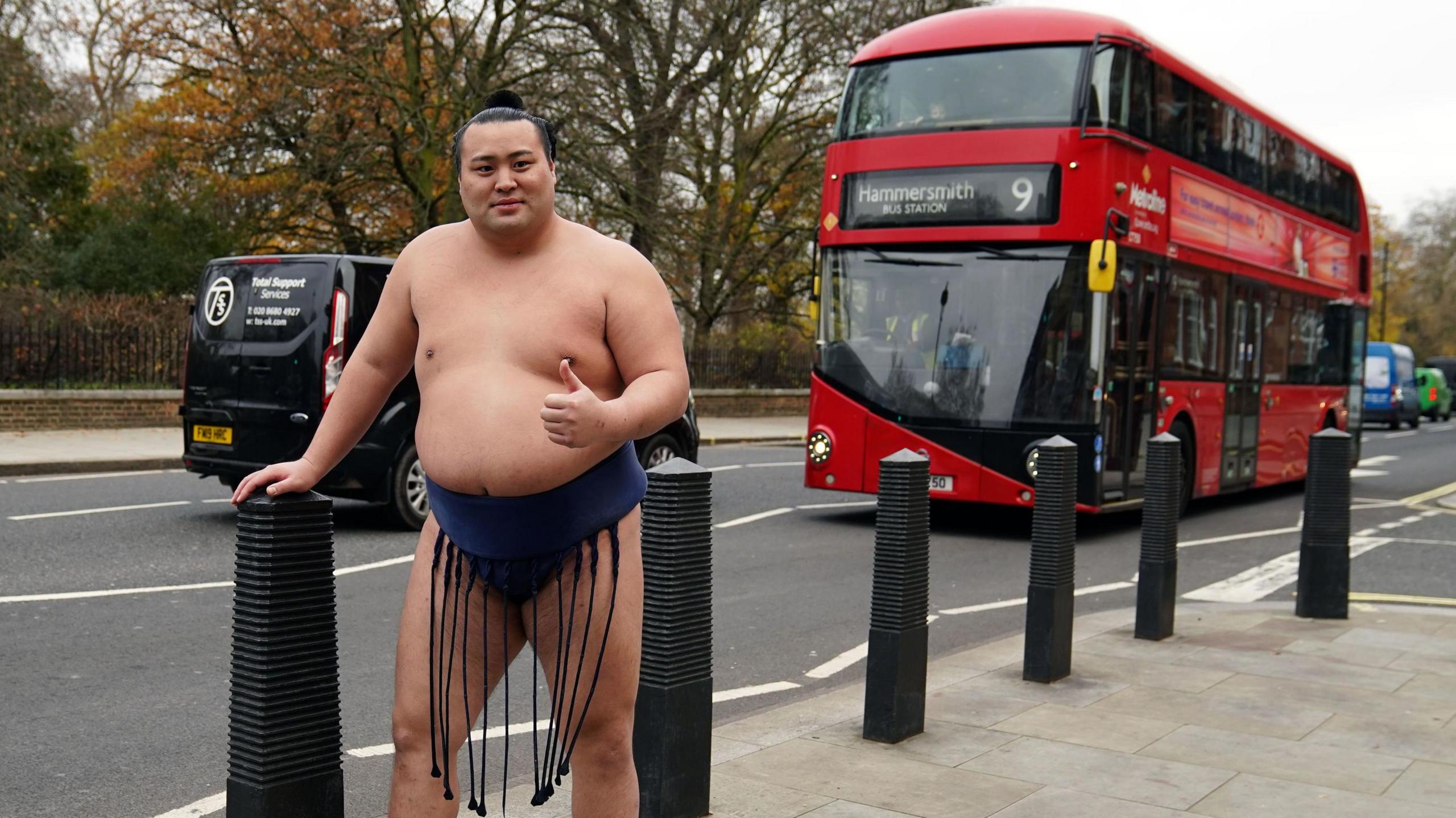 Sumo wrestler Kitanowaka Daisuke in a blue wrap standing in London with a red bus passing the back.