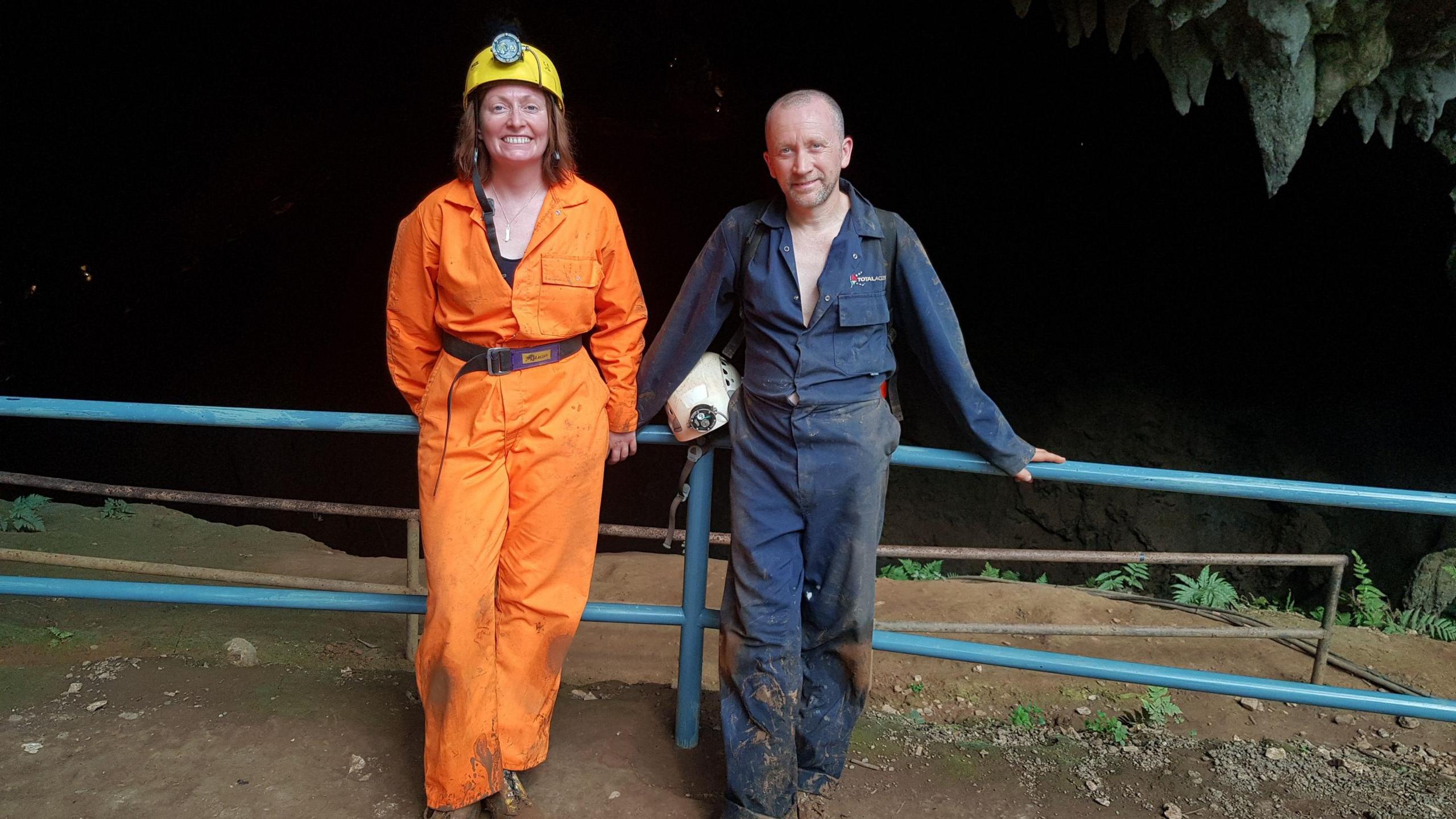 Mike Clayton in blue overalls holding a white helmet is pictured with his wife Emma in an orange overall jumpsuit. They are standing at the entrance to Tham Luang 