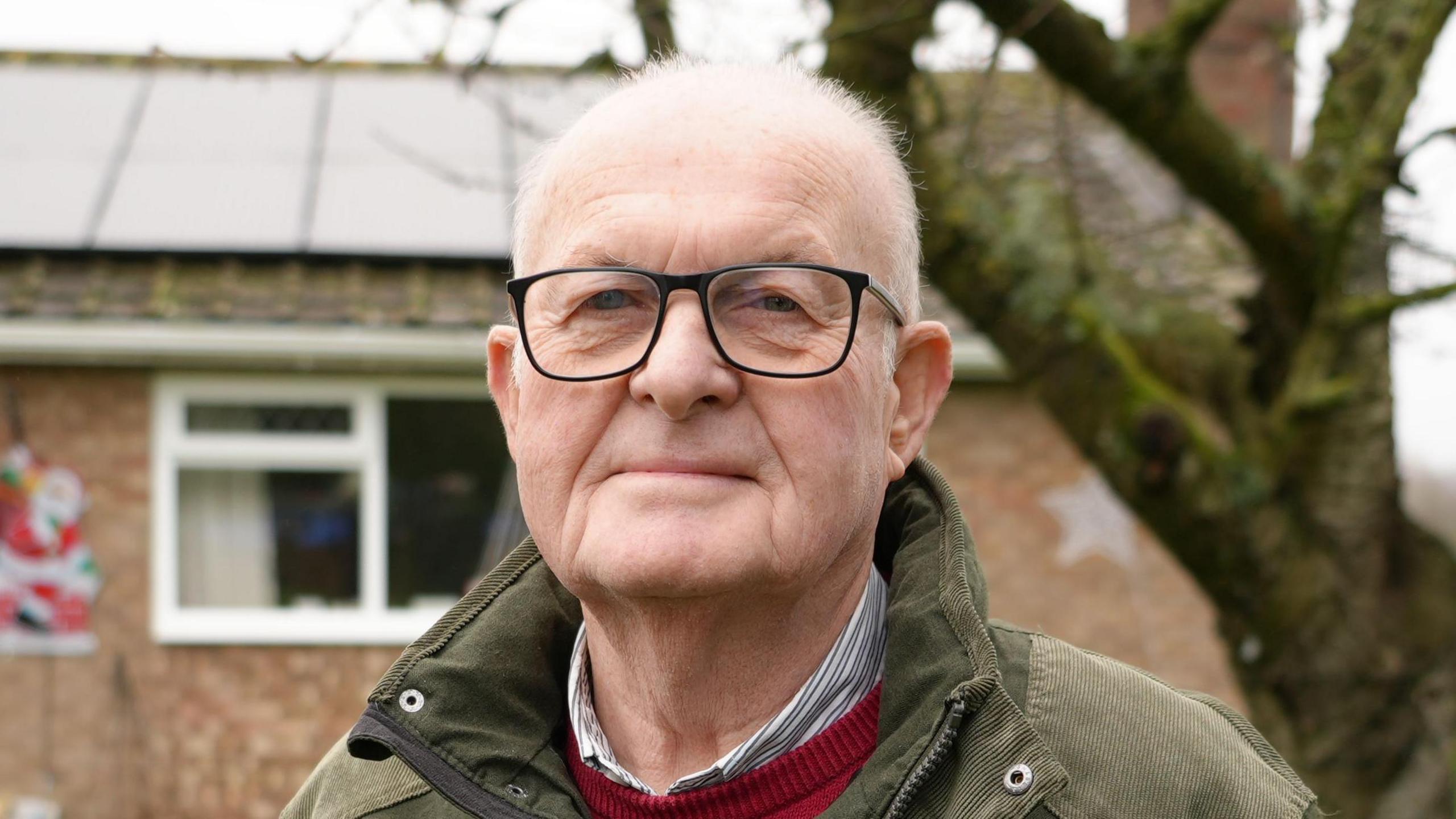 An elderly man smiles at the camera while standing in front of a home and tree. He is wearing dark glasses, a green coat, with a red jumper and shirt on underneath. 