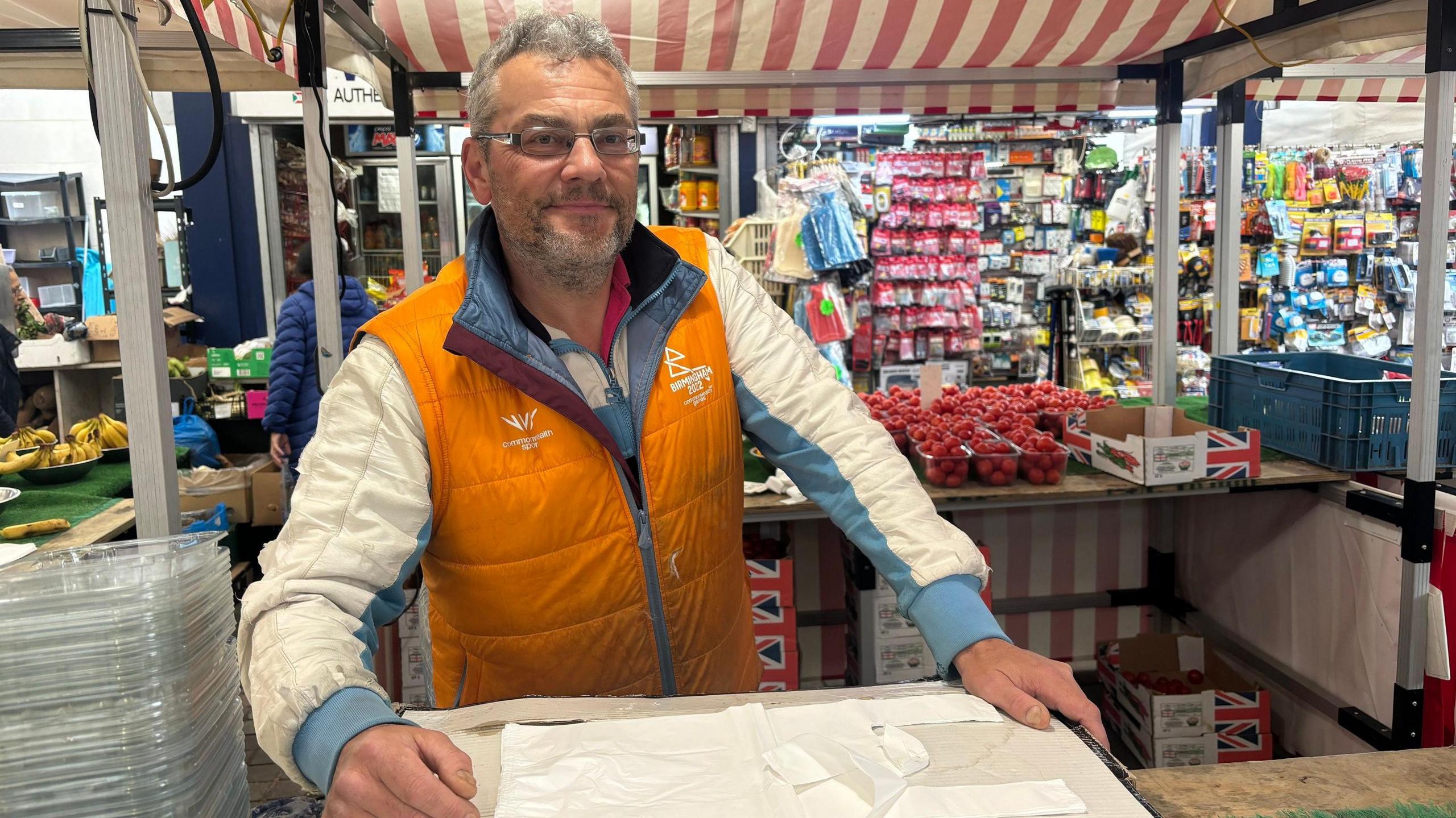 A market trader with an orange bodywarmer over a blue and white jacket stood at the serving area of his stall.