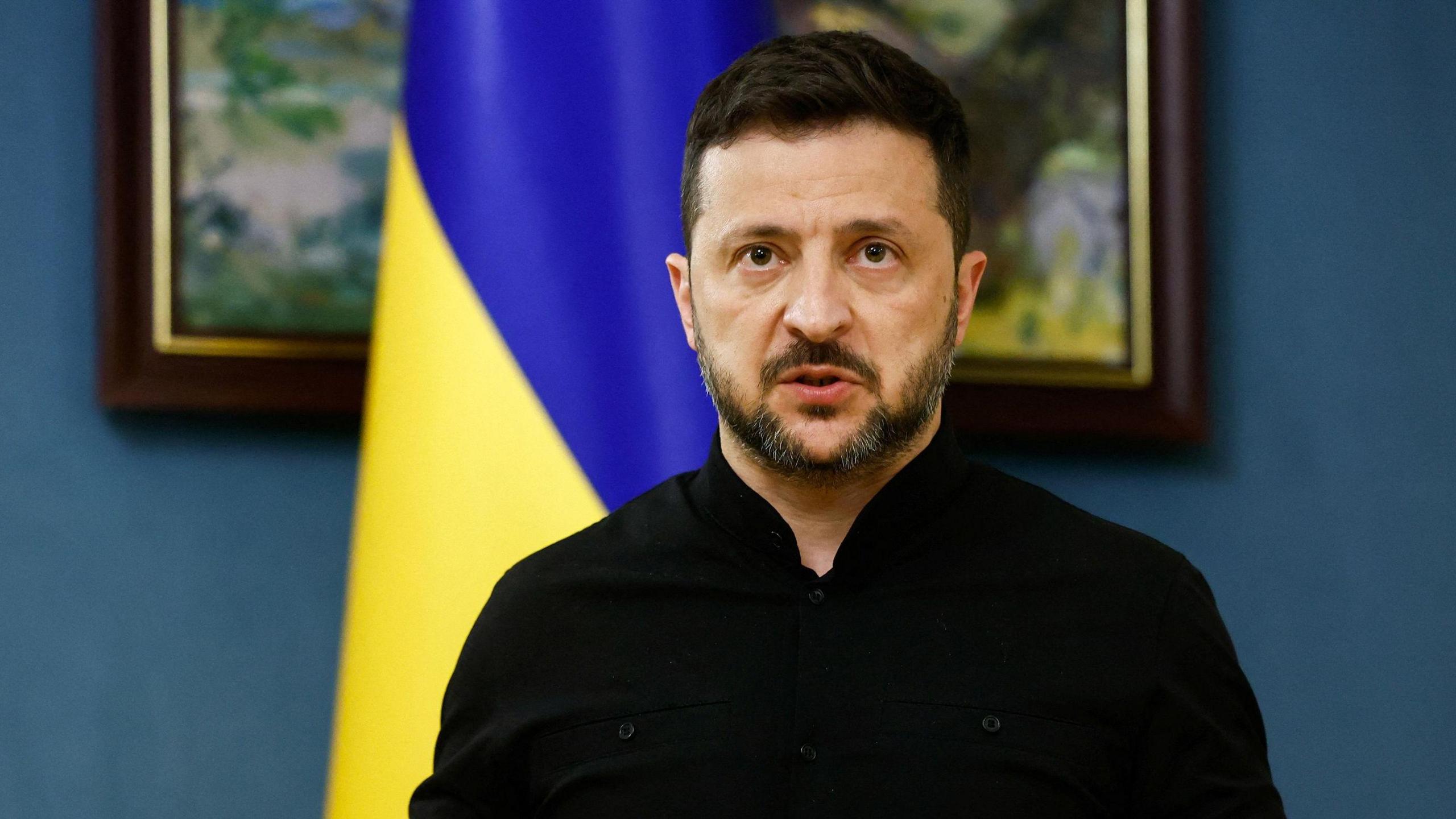 Zelensky speaking during press briefing following calls with Trump. behind him is a blue wall, a picture frame and a Ukrainian flag