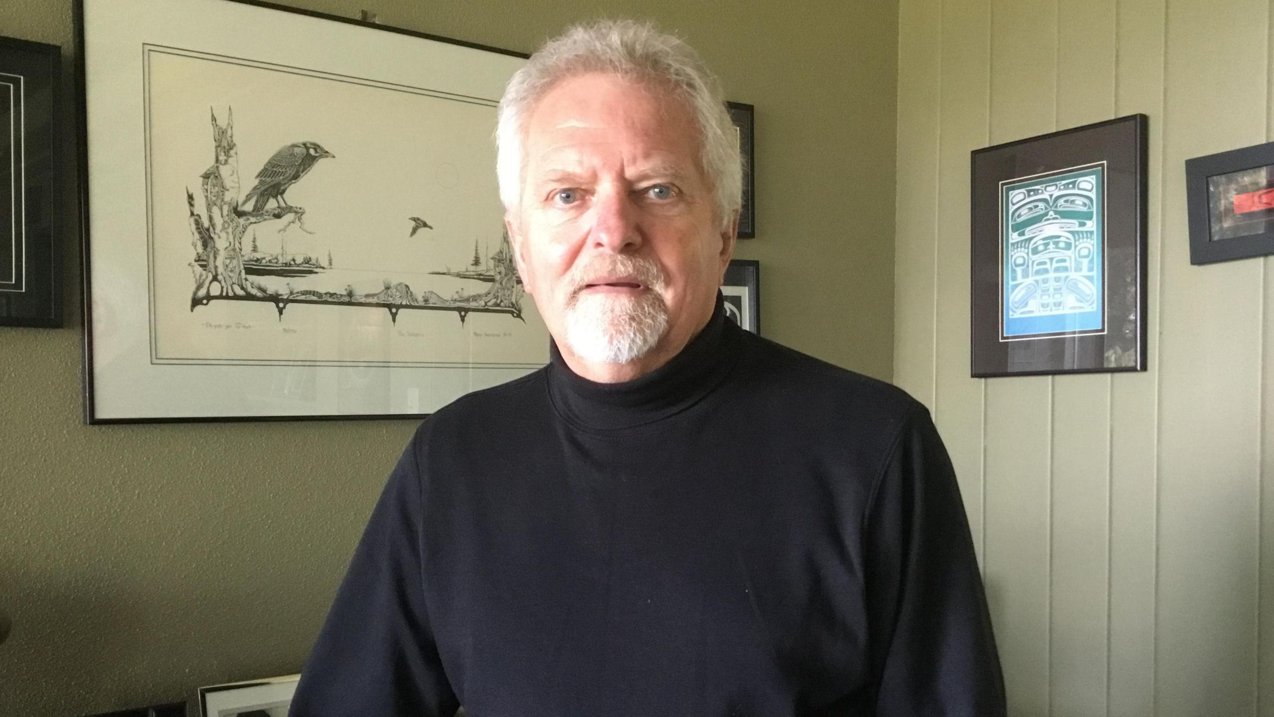 A portrait of Ian Andexer, 72, standing in his living room wearing a black turtleneck jumper. He has blue eyes and white short hair and beard.