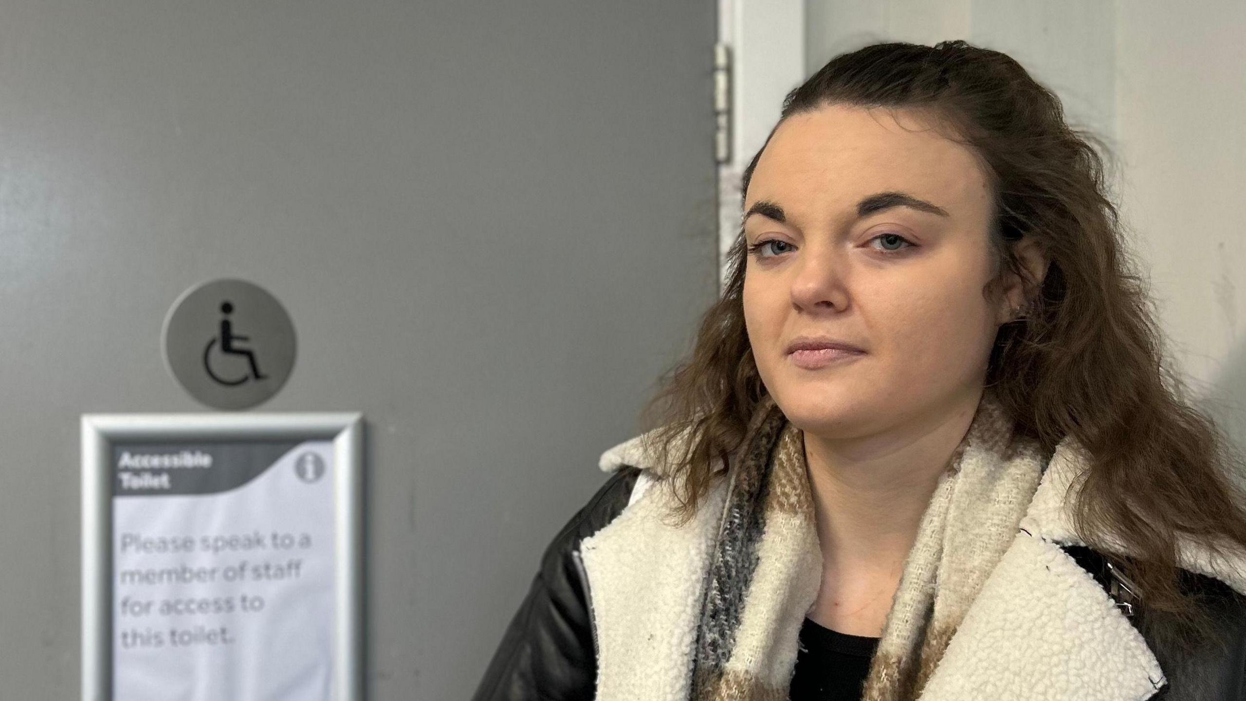 A young girl stands in front of a disabled toilet. She has dark brown hair, is wearing a brown chequered scarf and a black jacket with a white fur trim. A sign on the door reads: "Accessible toilet - please speak to a member of staff for access to this toilet".