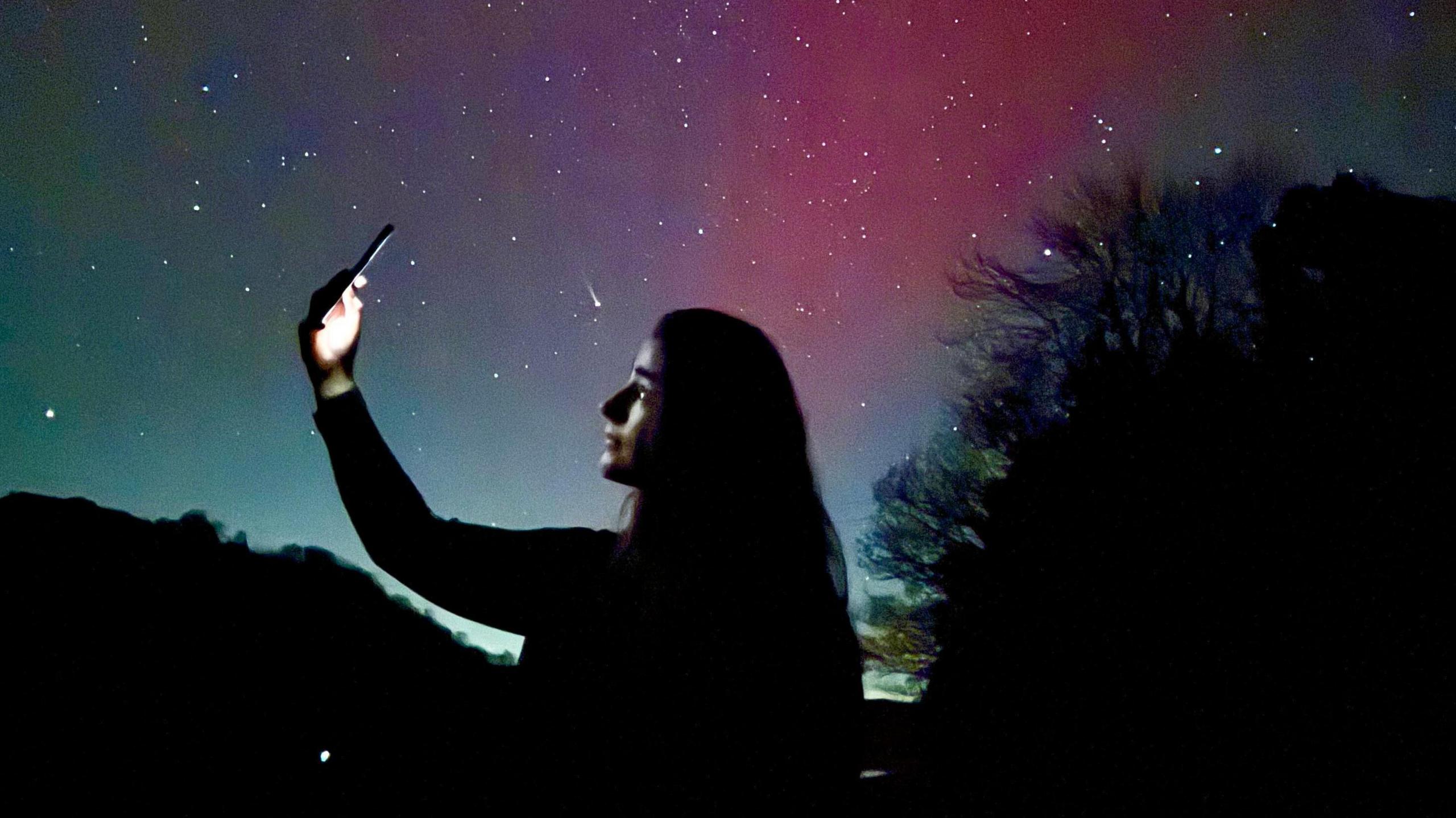 Woman pictured taking selfie seen from the side. She has long hair and the outline of the body and face are dark against the green and pink sky behind, and a light shines from her phone screen.