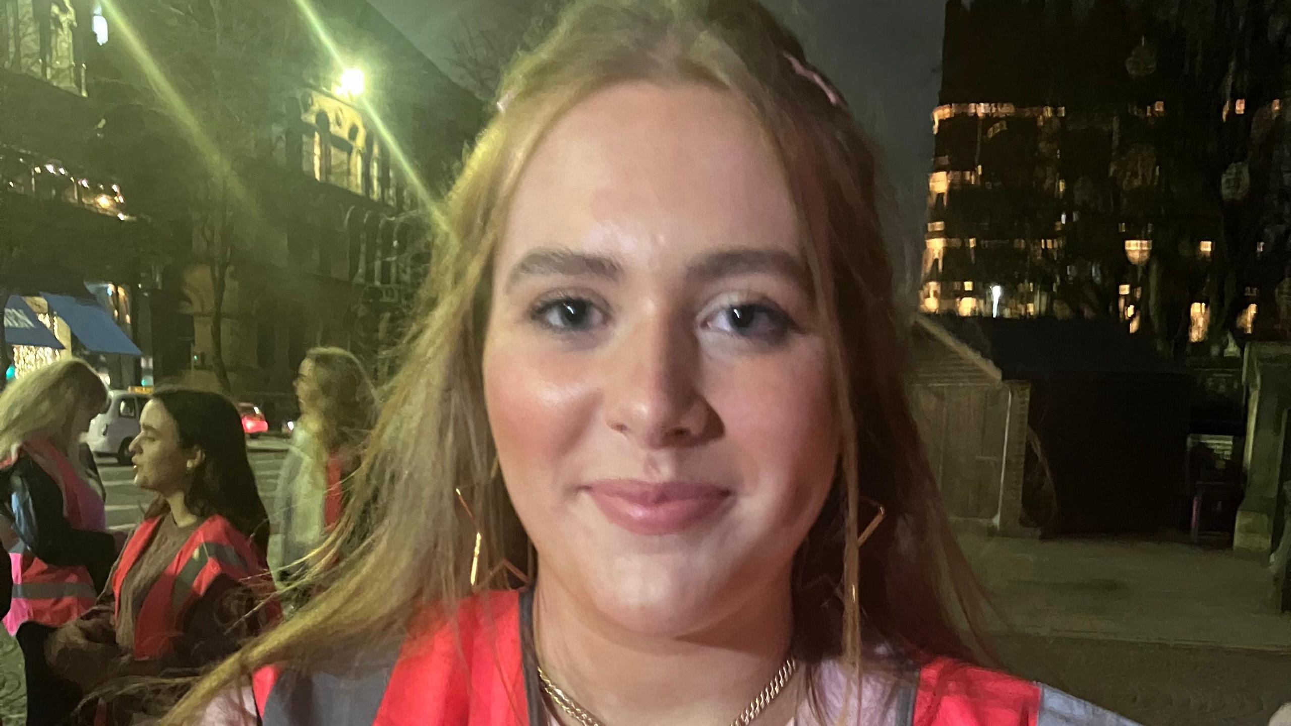 A girl with brown hair and green eyes wears a pink hi-viz over a pink jumper. She has on a gold necklace and gold star earings