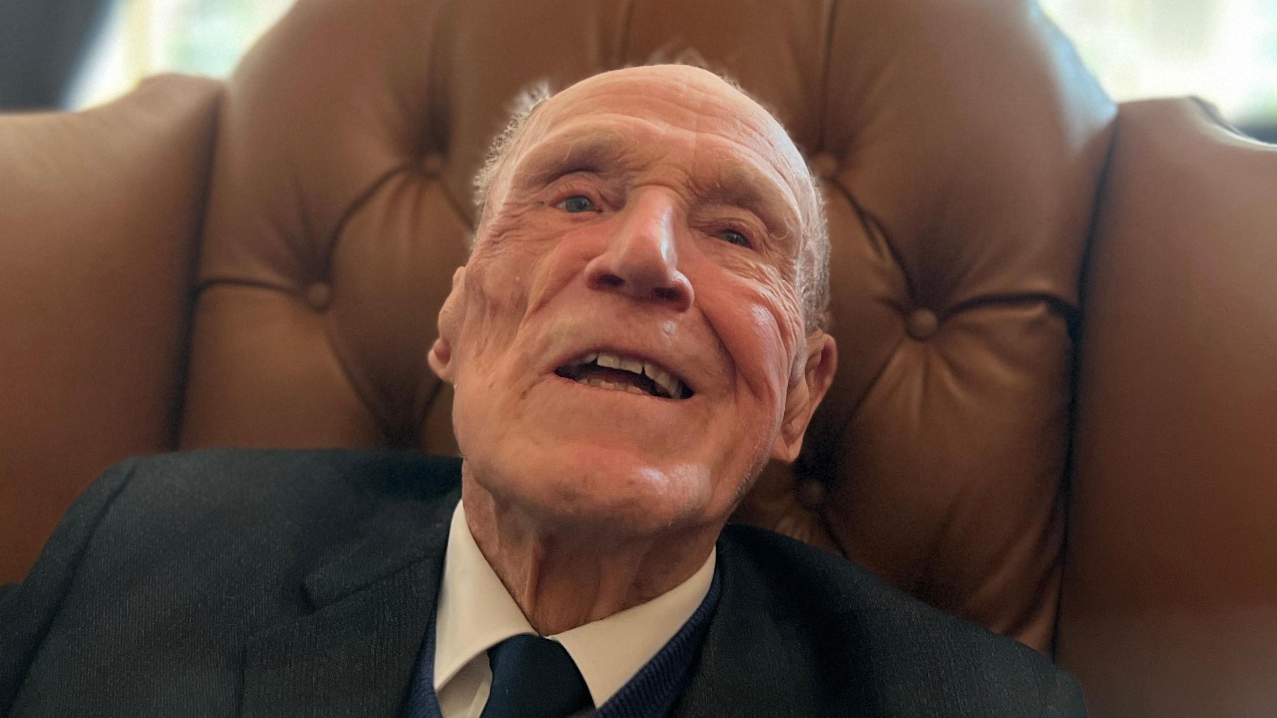 A close up photograph of Len Chivers, wearing a suit and tie while sat on a brown leather chair.