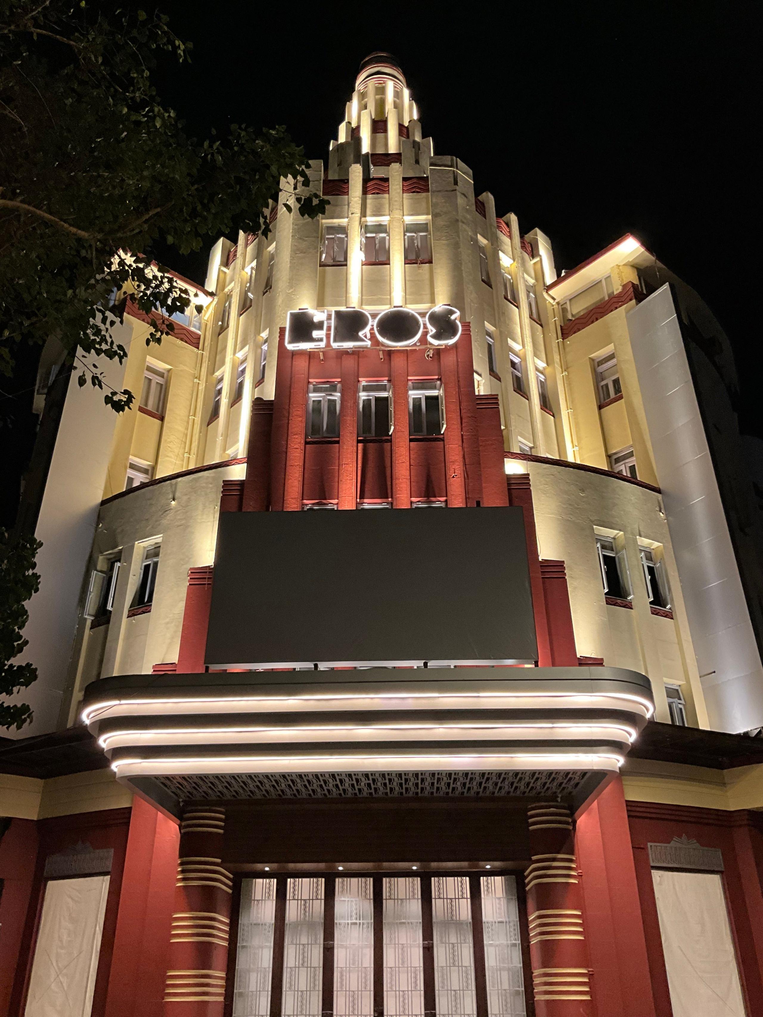 A night-time image of a lit up Eros Cinema, a Unesco-designated Art Deco theatre at Churchgate in Mumbai. The multi-storey building, which has a spherical tower at its top, is painted cream and partially faced with red sandstone. 