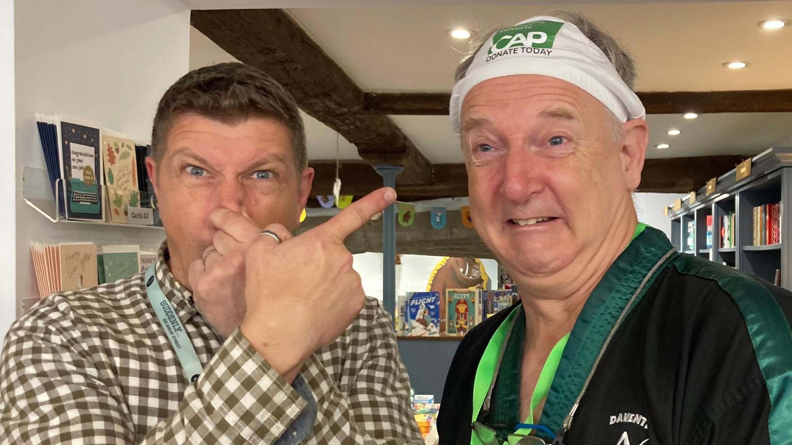 Tom Welch with short dark hair wearing a white and brown checked shirt with one hand over his nose and the other pointing at Mark Robson with white pants on his head with a green Christians Against Poverty logo.  They are standing in a bookshop.