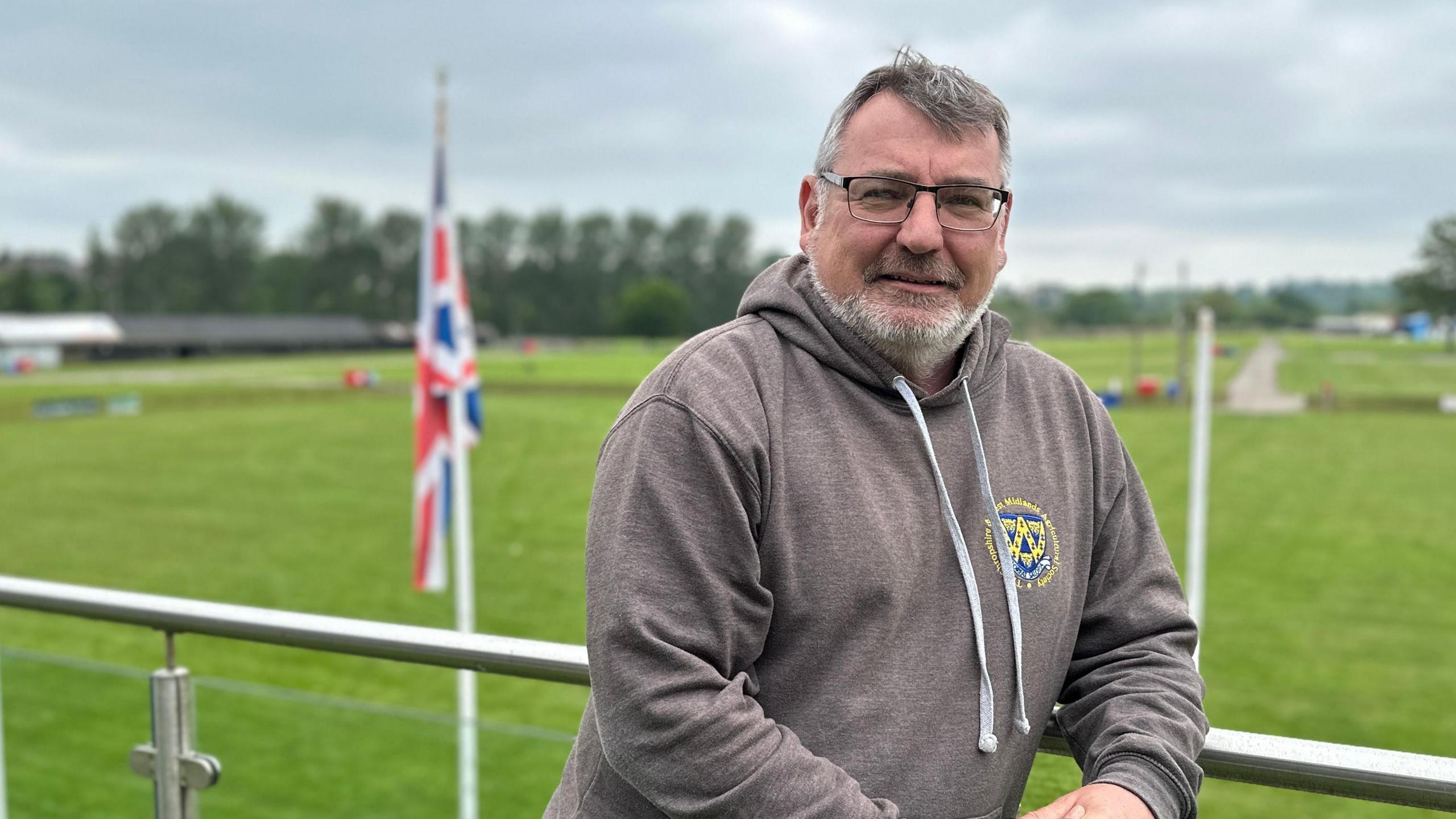 Ian Bebbington is in a grey hoodie. He has grey hair and facial hair and is wearing black glasses. He is leaning on a metal rail of a balcony, behind him is a stretch of green grass and a white flagpole with the union jack hanging from the top.