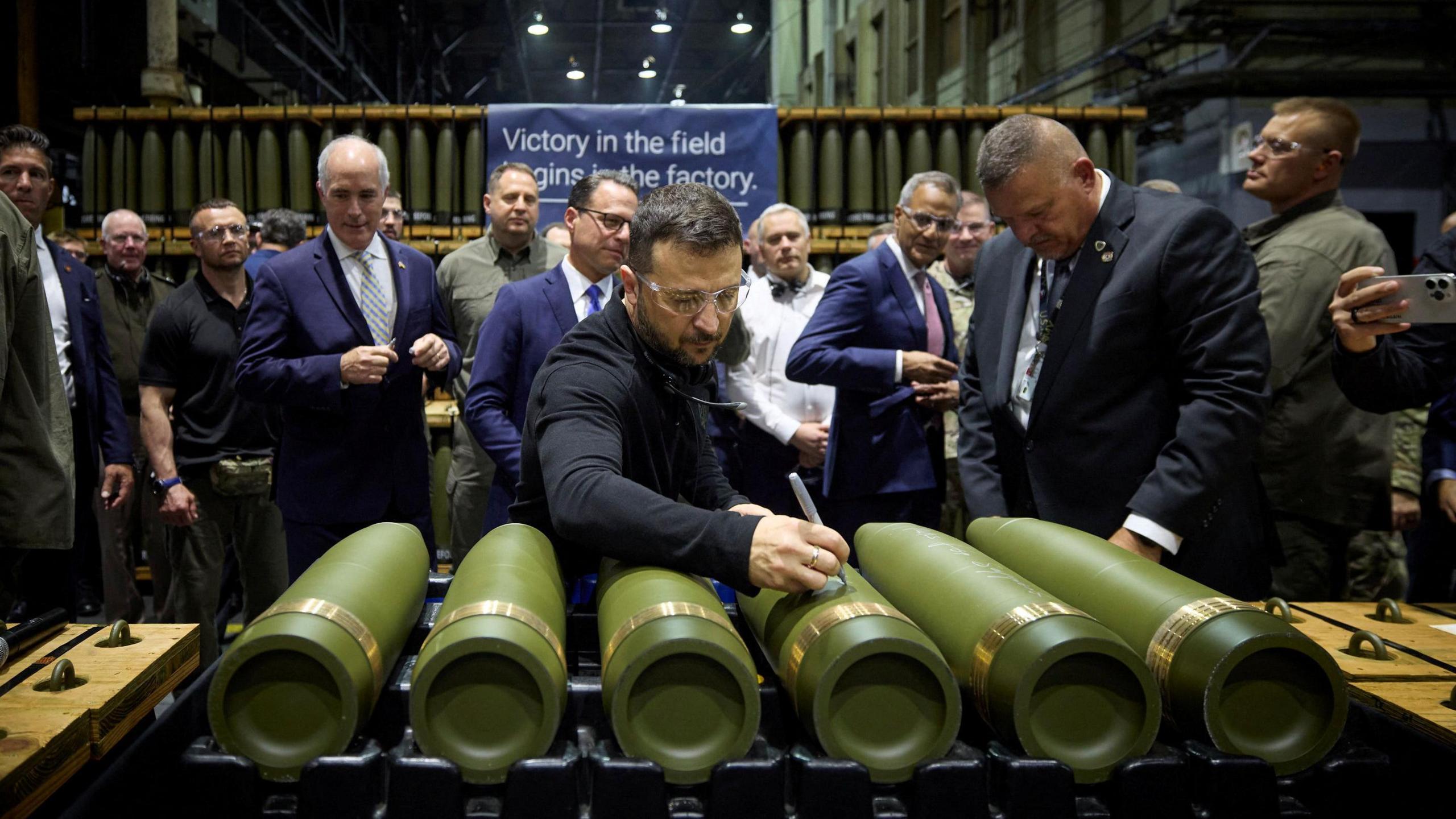 Flanked by other officials, Ukrainian President Volodymyr Zelensky writes with a pen on an artillery shell in a factory