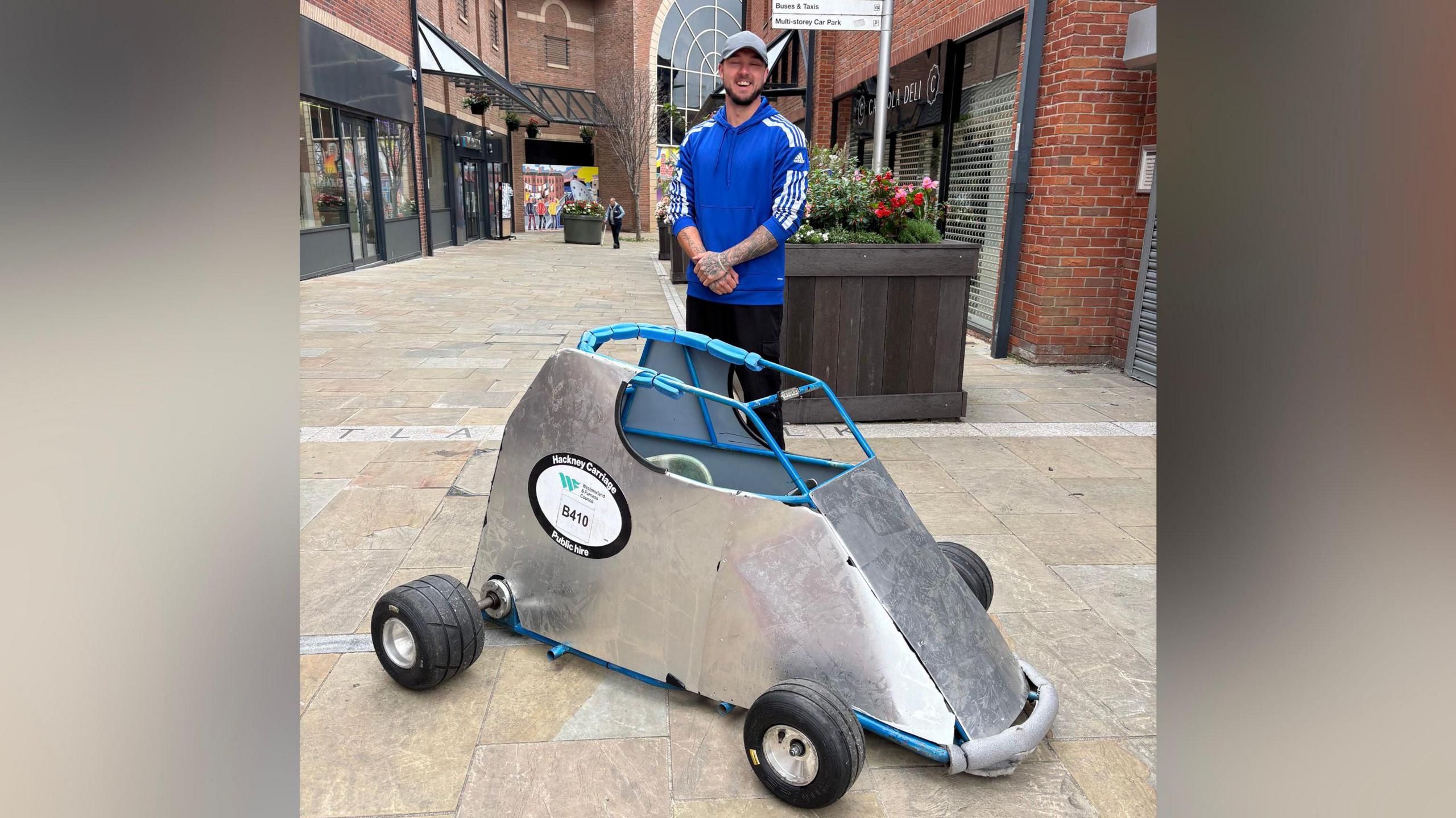 A man wearing a grey cap and blue top is standing, smiling beside the cart. It is covered in metal panels and has a sticker on it that reads: "Hackney Carriage".