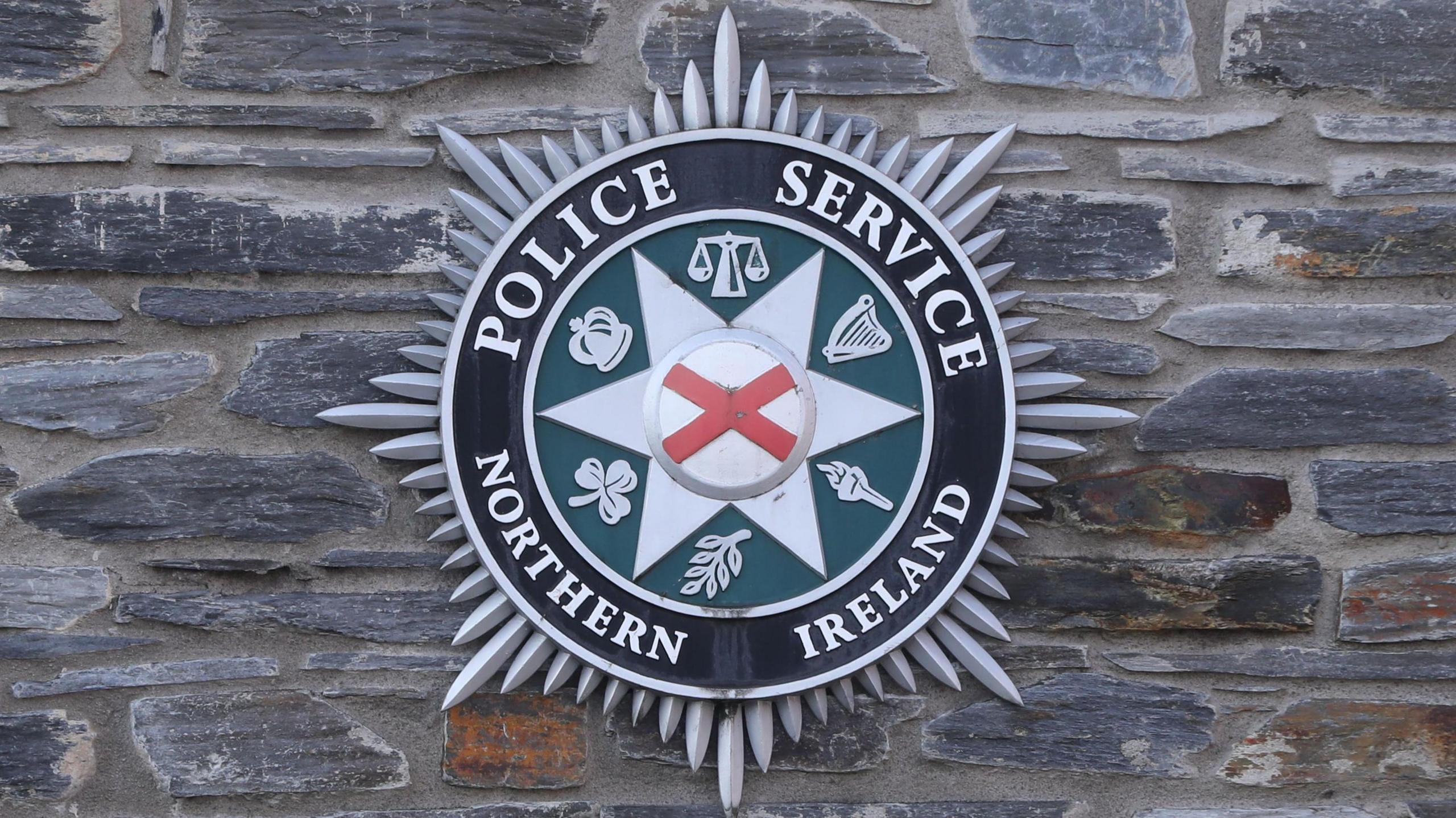 A large PSNI crest, displaying the Police Service of Northern Ireland, on a grey stone wall.