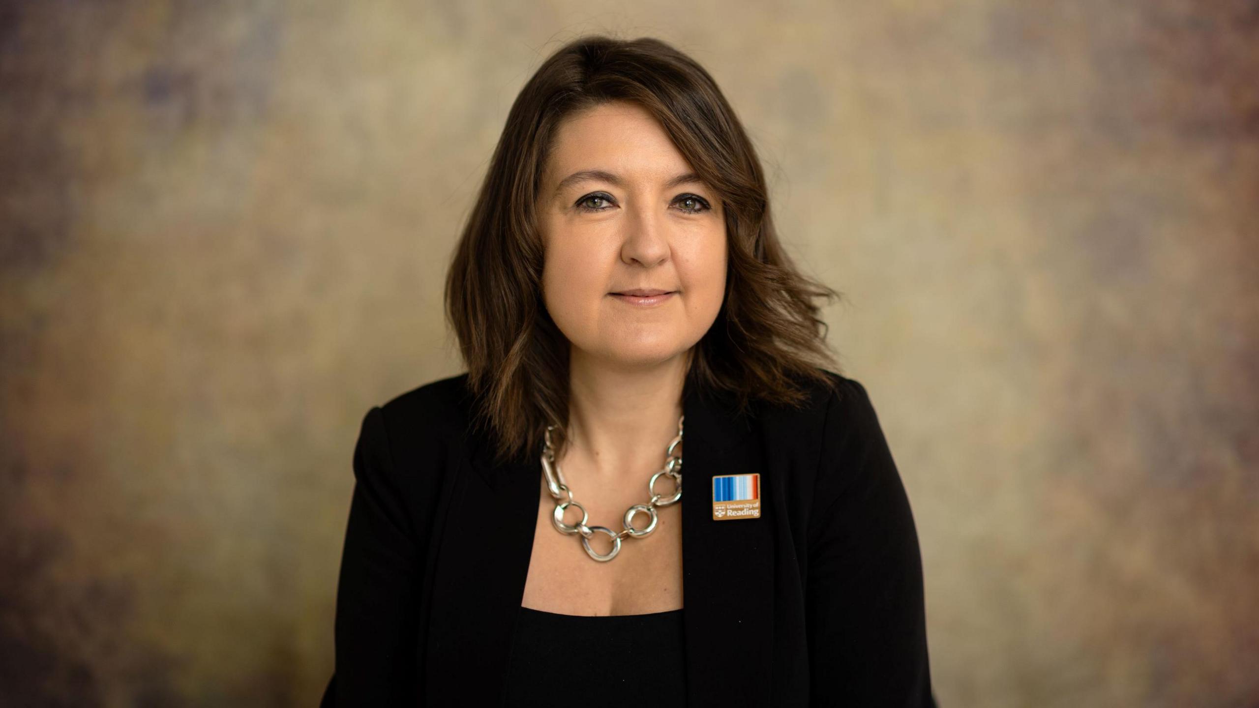 Caroline Baylon wearing a silver necklace and a black jacket with a black top underneath. She has a University of Reading logo pin badge attached to her jacket. 