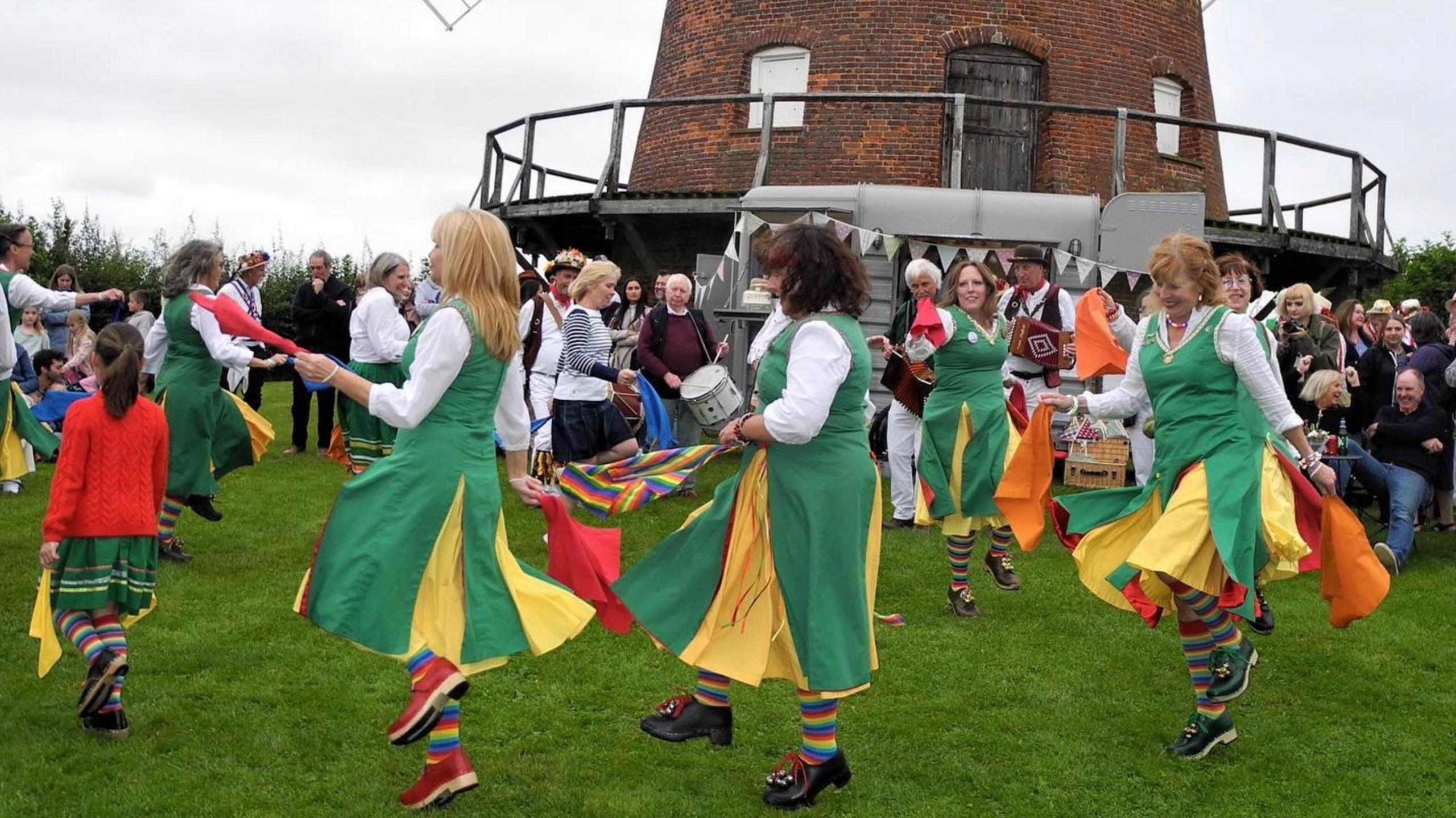 Female Morris dancers wearing white and green dance around the base of the windmill as a crowd watches on