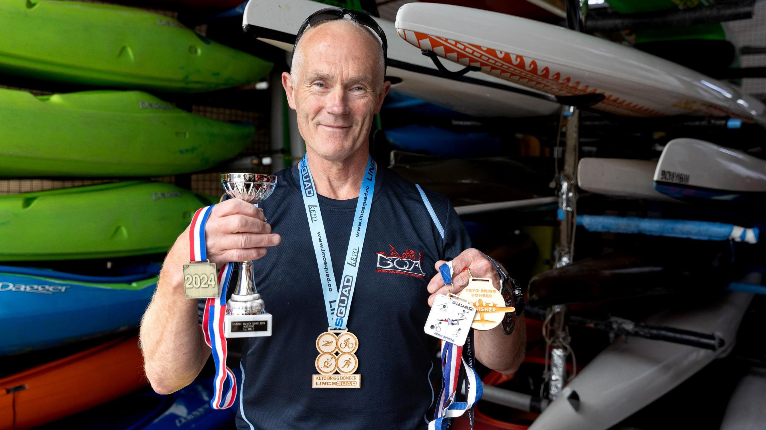 A man called Jules Taylor stands inside a shed lined with canoes and kayaks, holding various medals he won for becoming Quadrathlon World Champion for his age group. He is smiling and is wearing a dark blue T-shirt and has a pair of sunglasses perched on his head