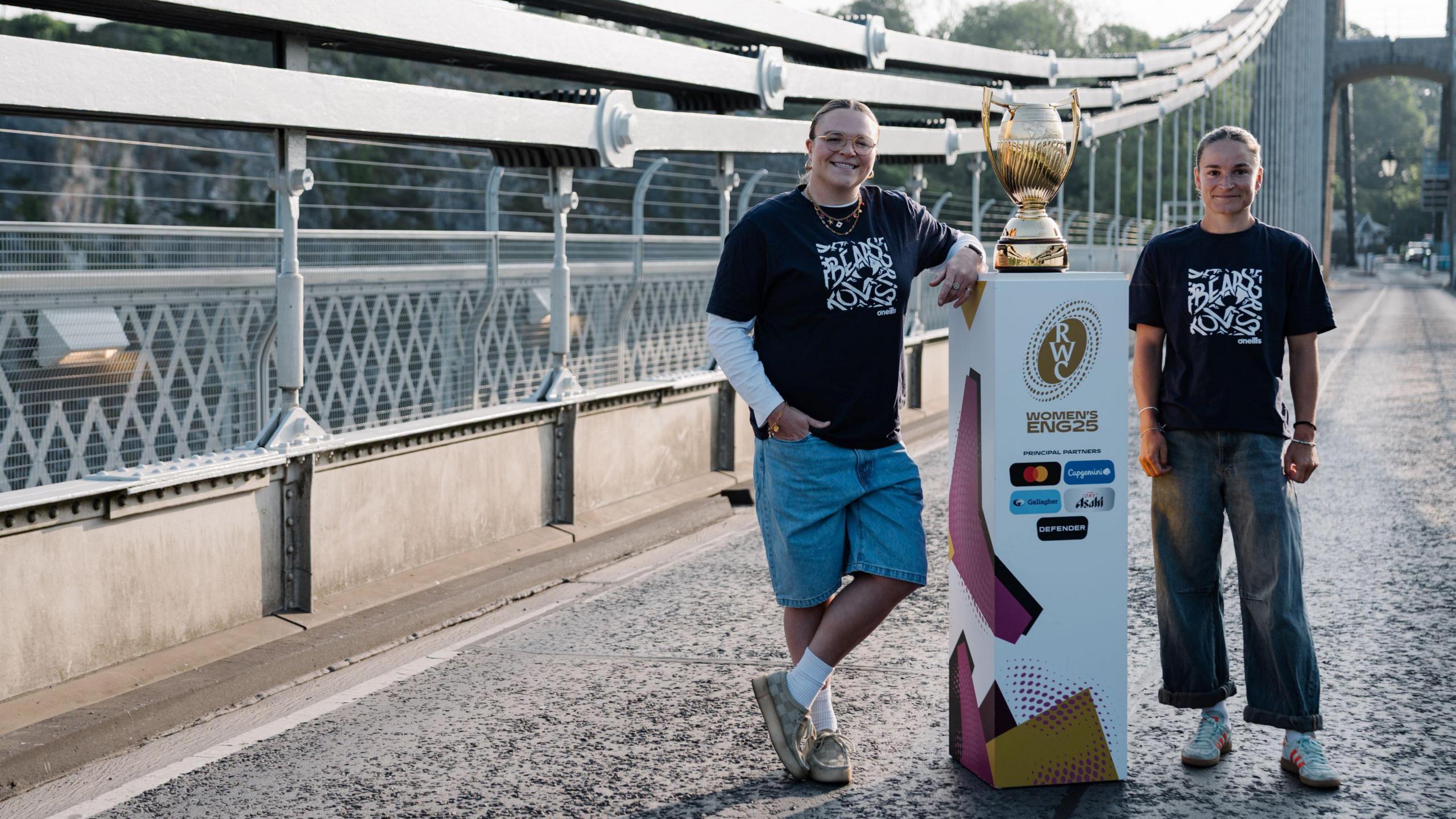Two young women, both wearing black T-shirts with white writing on them, stand either side of a colourful plinth which has the women's rugby world cup on it. They are on the Clifton Suspension Bridge in Bristol, and the large bridge supports are visible