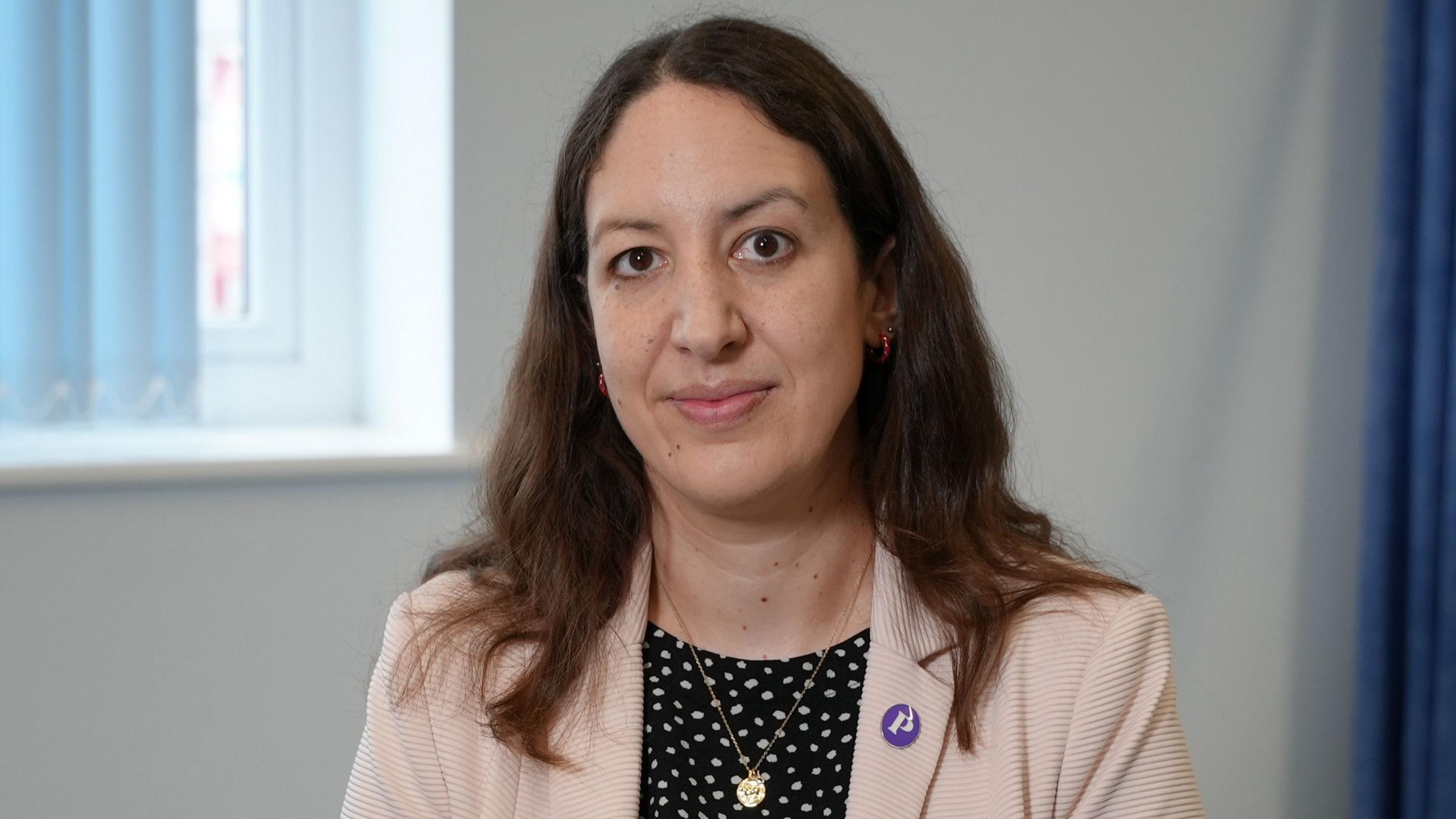 A woman with long dark, slightly wavy hair wearing a light pink jacket and a black top with white polka dots looks straight at the camera. She has a Pancreatic Cancer UK badge on her lapel and is also wearing a small gold medallion necklace. The wall behind her is white. There is a partial window visible to her left and a long blue curtain to her right.