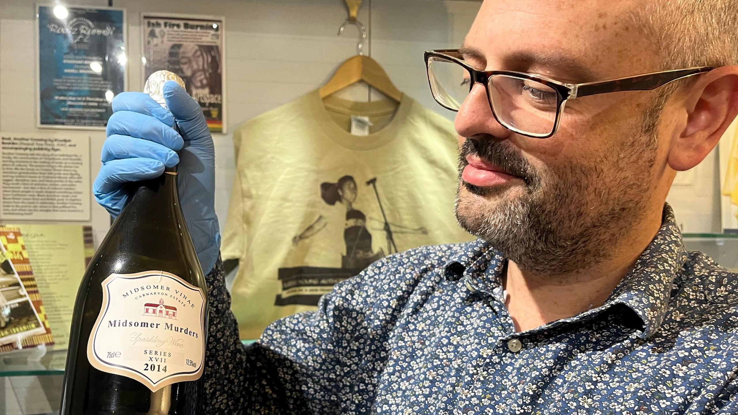 A man with short dark beard wearing a floral blue shirt holds a wine bottle. 