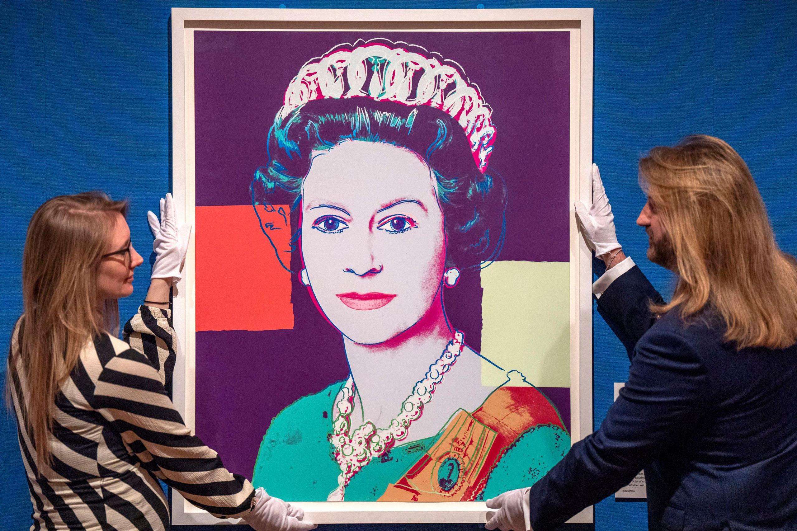 A colourful screenprint of the late Queen. She is wearing a green dress with orange sash. Two curators are holding either side of the picture. One is a man with long brown hair and a brown beard. The other is a woman who is wearing a black and white stripped top. She has long brown hair and is wearing glasses. They both have white gloves on.