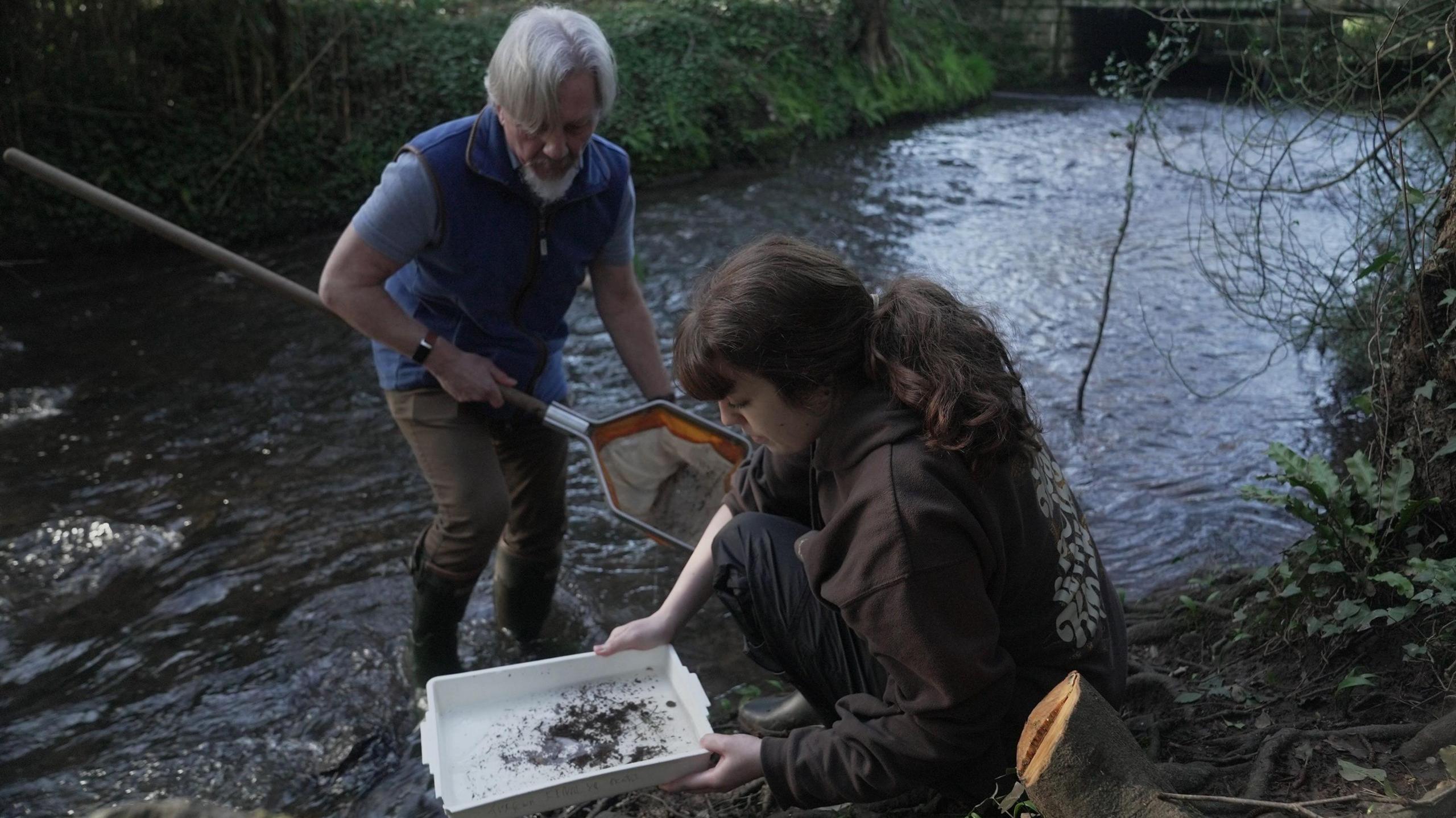 Ymchwilwyr o Brifysgol Caerdydd yn samplo dwr yn Nant Lleucu, Caerdydd. Mae'r Athro Steve Ormerod yn sefyll yn y dwr gyda rhwyd a Molly Hadley ar lan yr afon yn dal padell wen. 