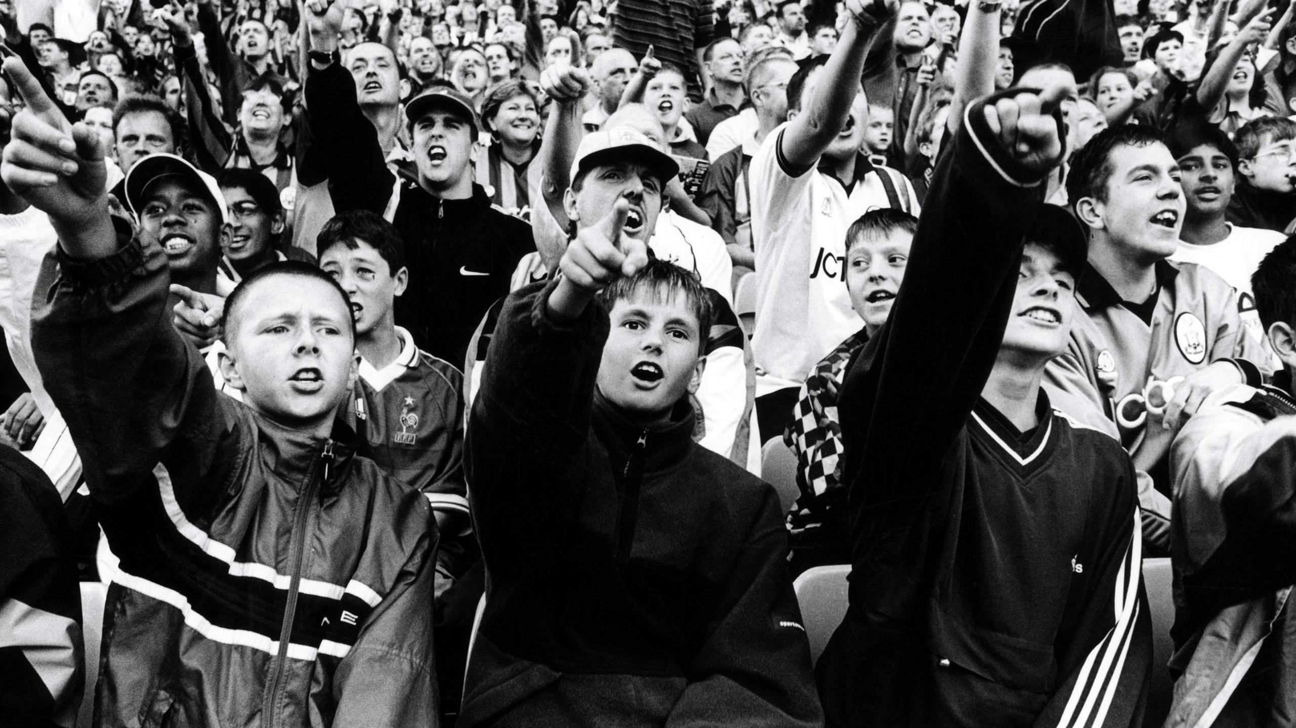 A black-and-white photo of a football crowd with three teenage boys in tracksuits at the front pointing into the air.