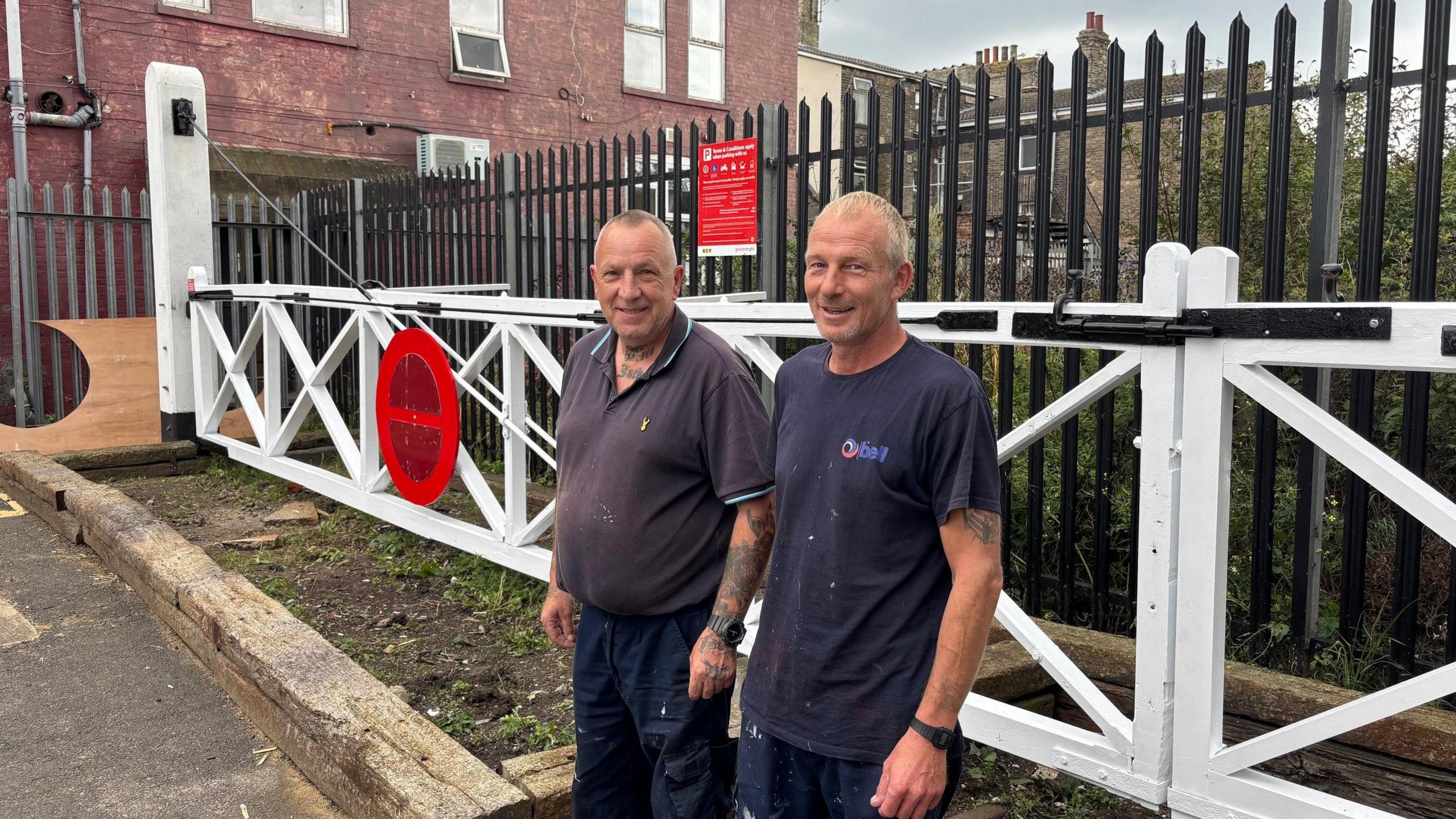 Two men wearing navy-coloured clothing and standing in front of the railway gates. They are both looking at the camera and smiling.