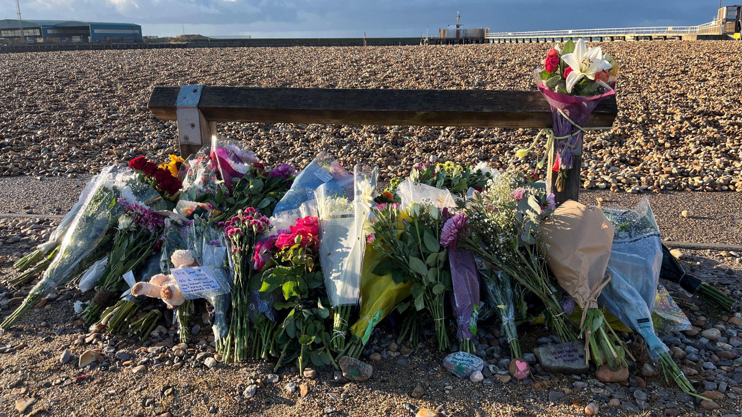 A pile of flowers left by a wooden post next to a shingle beach.