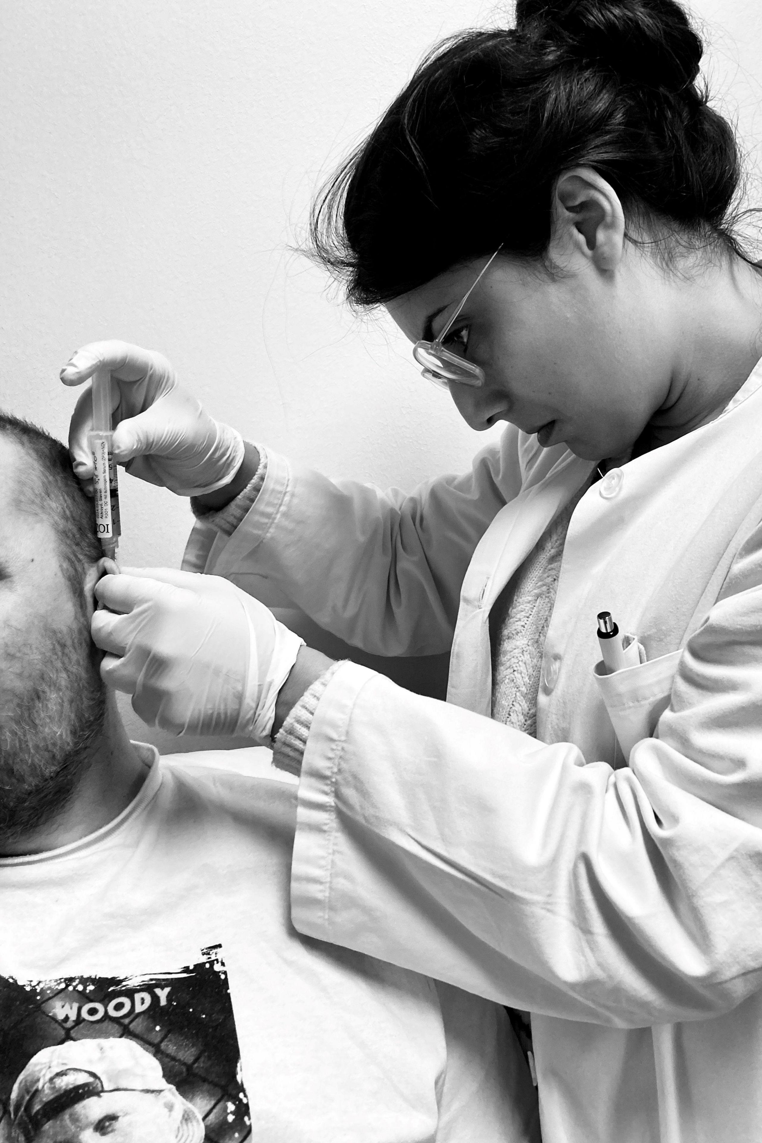 A female medical professional in a lab coat and gloves administers an injection behind a Steve Ackroyd's ear.