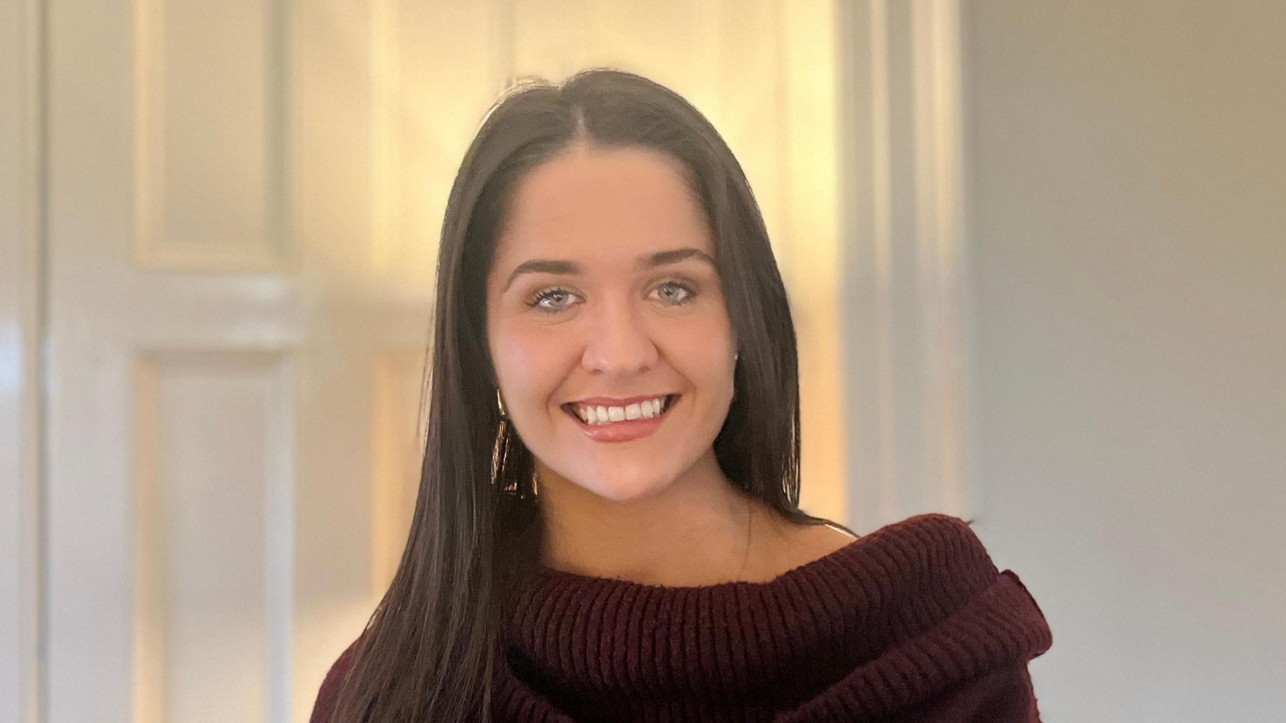 Suzy Spence. She has long dark brown hair, blue eyes and smiling directly into the camera and wearing a dark red jumper.