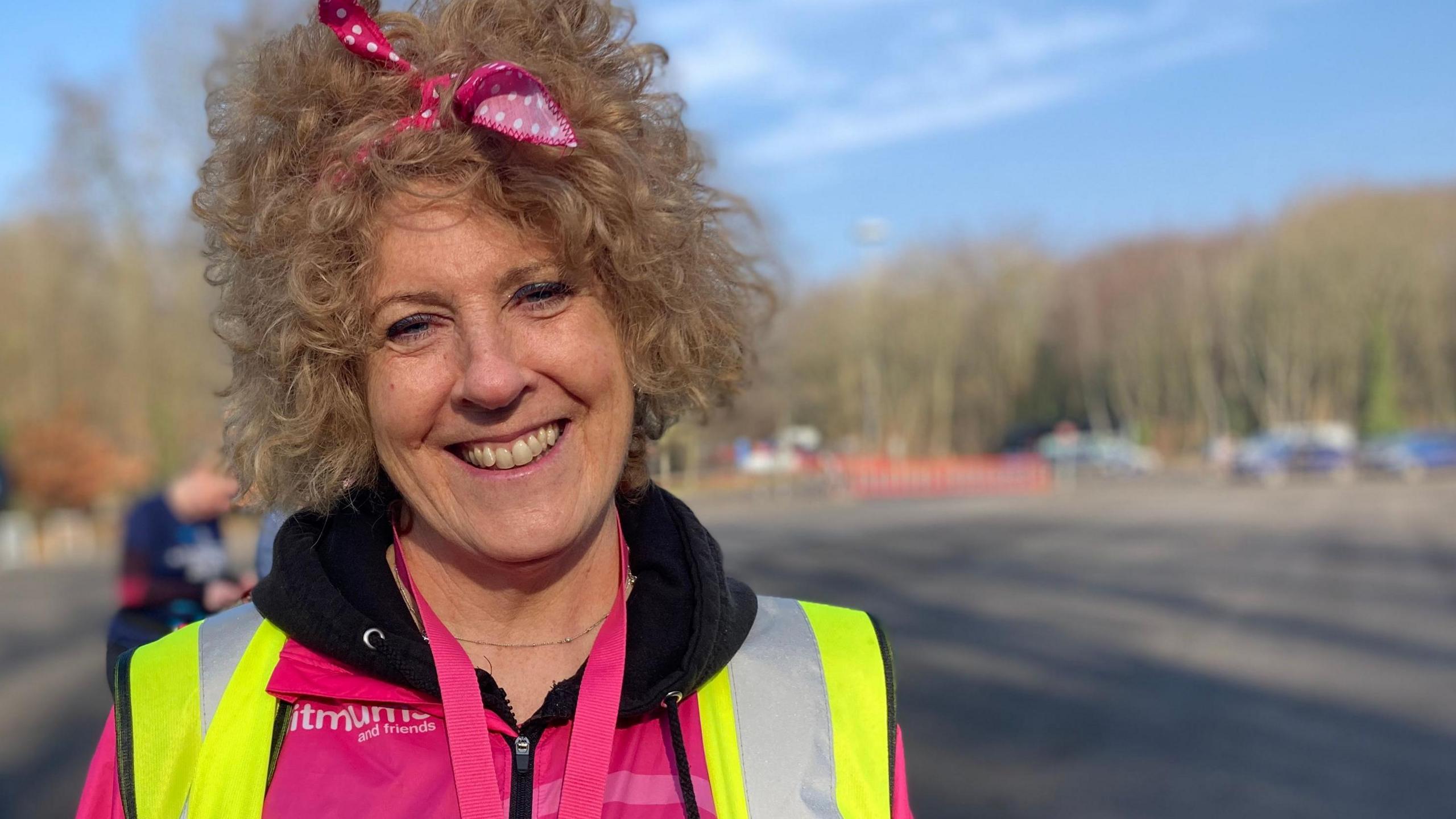 A woman smiles while standing in a large car park. She has frizzy light-brown hair and there is a pink bow in it. She is wearing a pink shirt with a yellow hi-vis vest over the top of it.